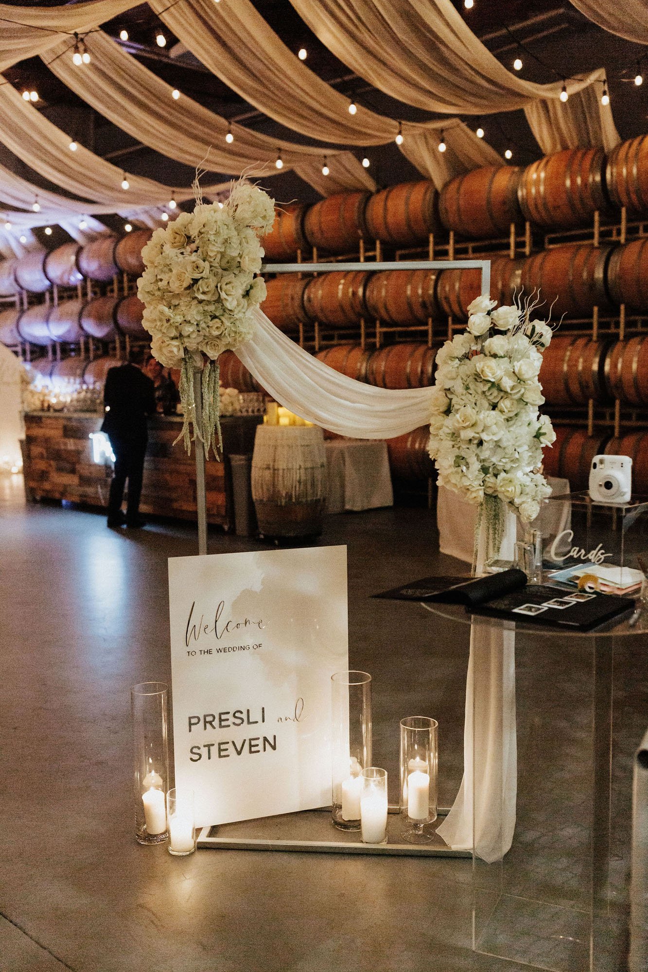  Photograph of a welcome table at a vineyard wedding in Washington. There is a floral installation with white roses, hydrangeas, and draped silk. There is a sign on the ground that reads “Welcome to the wedding of Presli and Steven” that is surrounde