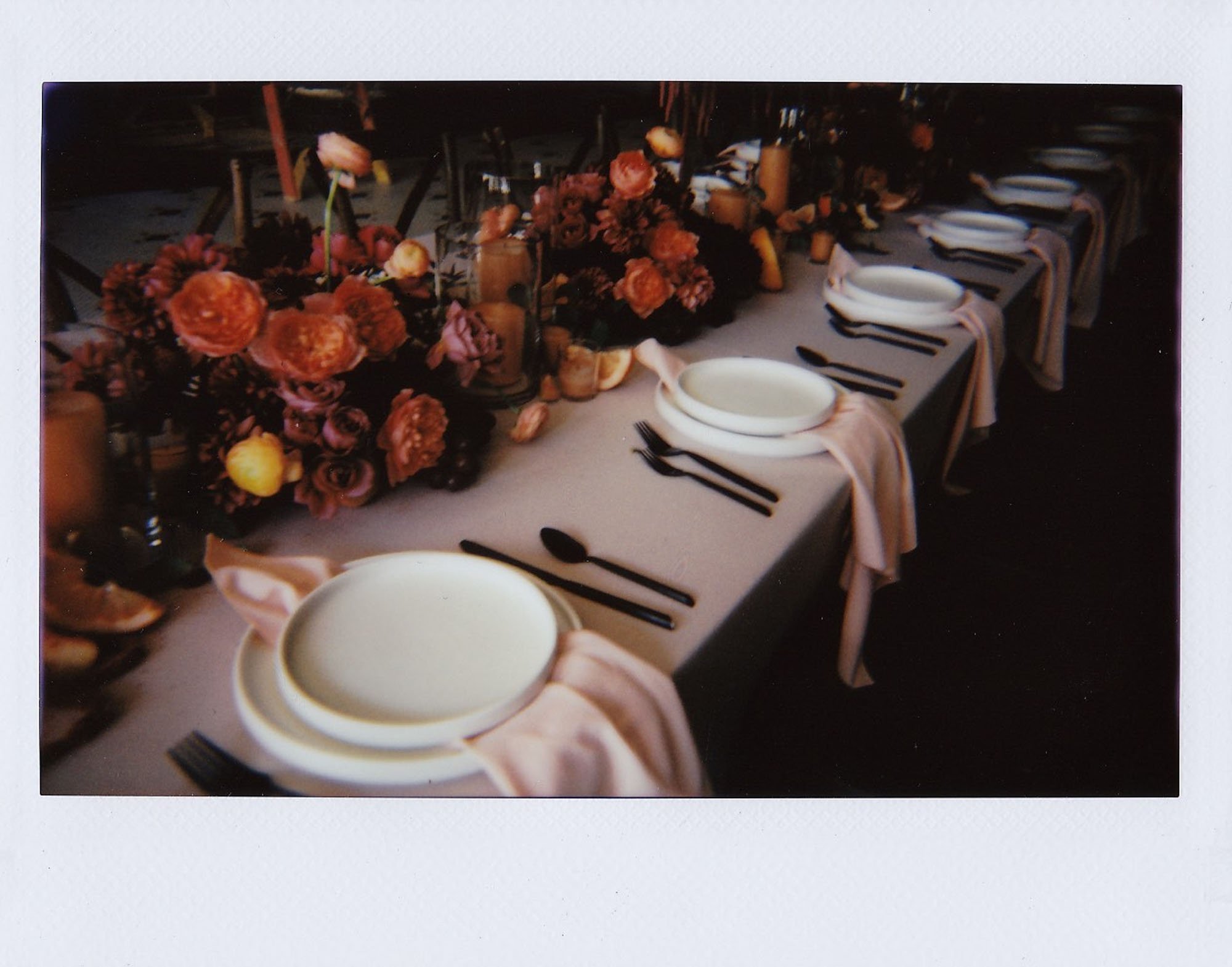  polaroid photo of a colorful and modern wedding table setting, with black flatware, white dinner plates, blush silk napkins, and lush floral centerpieces of burgundy, pink, orange, and red. 