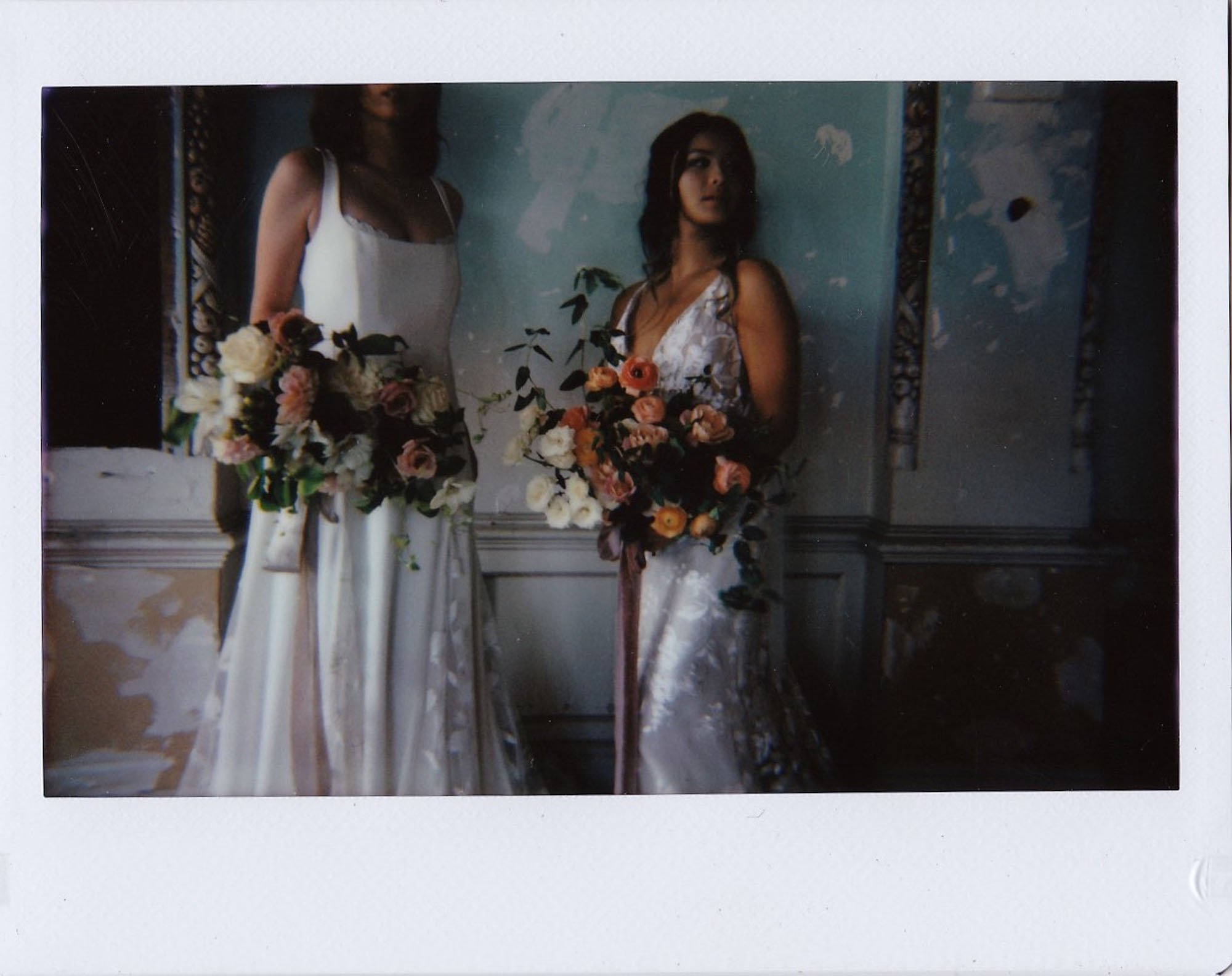  two brides standing side by side, one is wearing Alexandra Grecco Sienne wedding dress, and the other is wearing Made With love Elsie wedding dress. Both brides are holding large bridal bouquets with long romantic silk ribbons. 