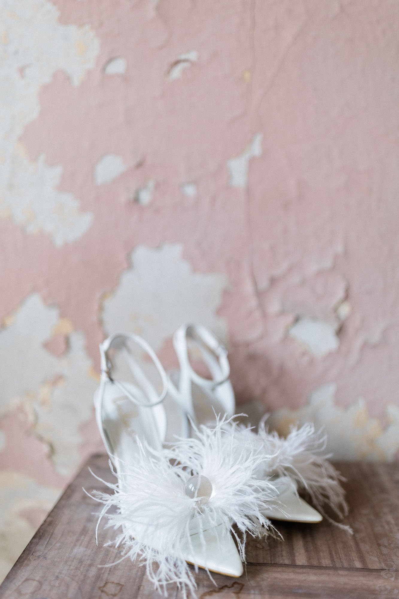  a lovely pair of white bridal shoes. they are high heels with open pointed toes and large white feather accents. they are on a wood table in front of a wall with peeling pink paint. 