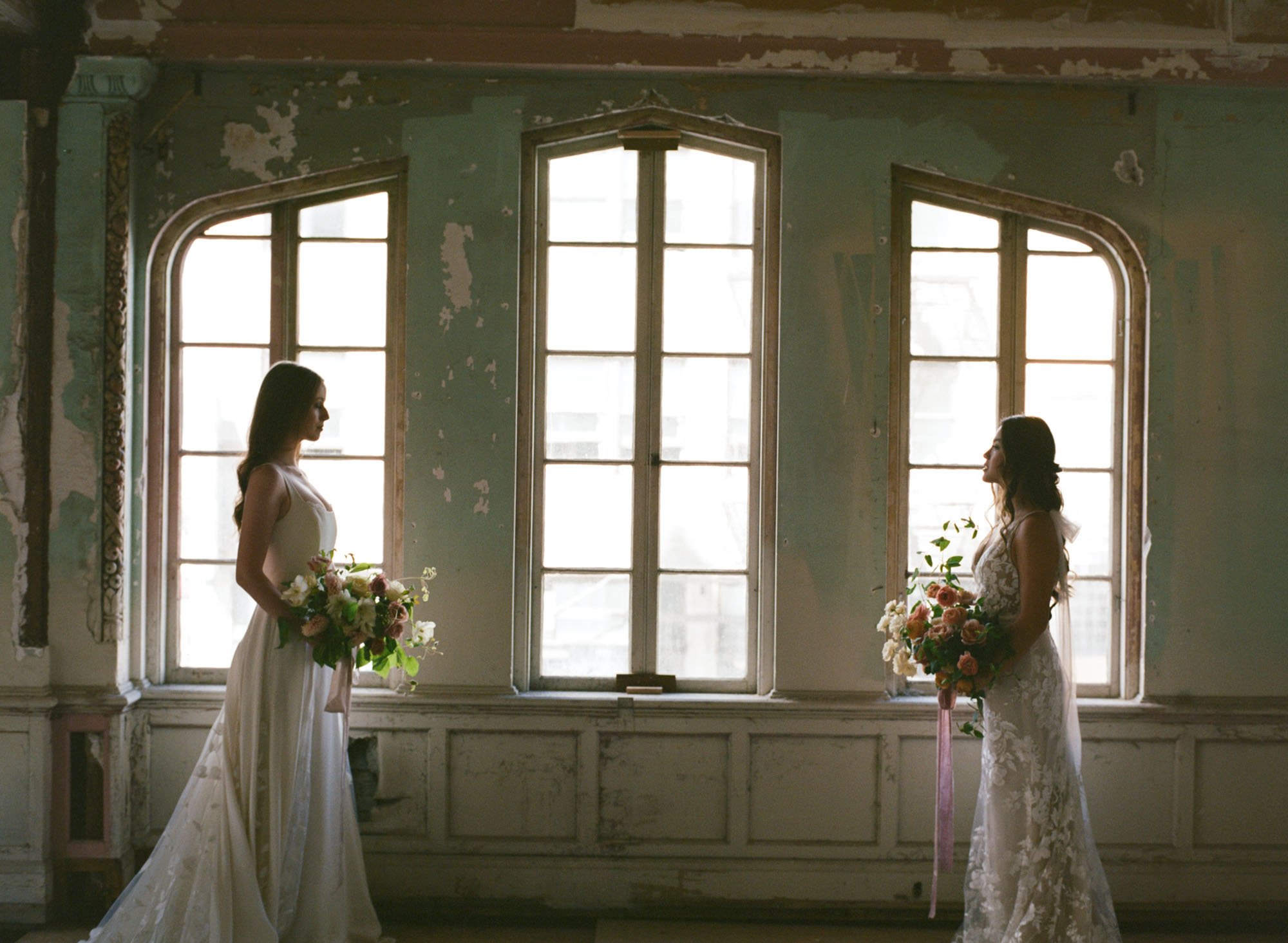  two brides standing in front of old bay windows in an abandoned hotel. one is wearing Made With Love Elsie wedding dress, and the other is wearing Alexandre Grecco Sienne wedding dress. they are each holding a bridal bouquet. 