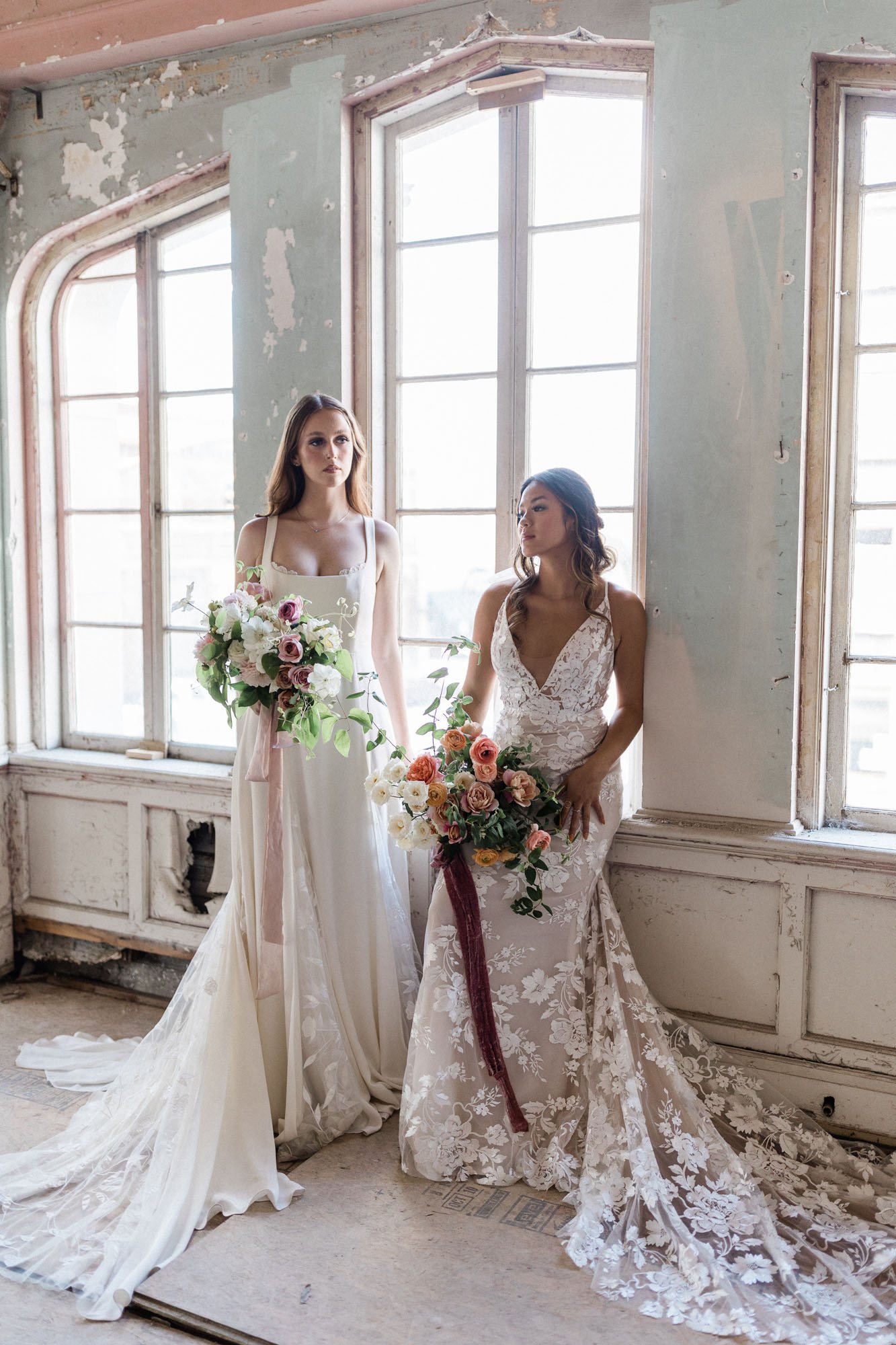  Two brides sitting in a window ledge of an old abandoned hotel in Oregon. One is wearing Alexandra Grecco Sienne wedding dress and the other is wearing Made With Love Elsie wedding dress. They are each holding large bridal bouquets with silk ribbons