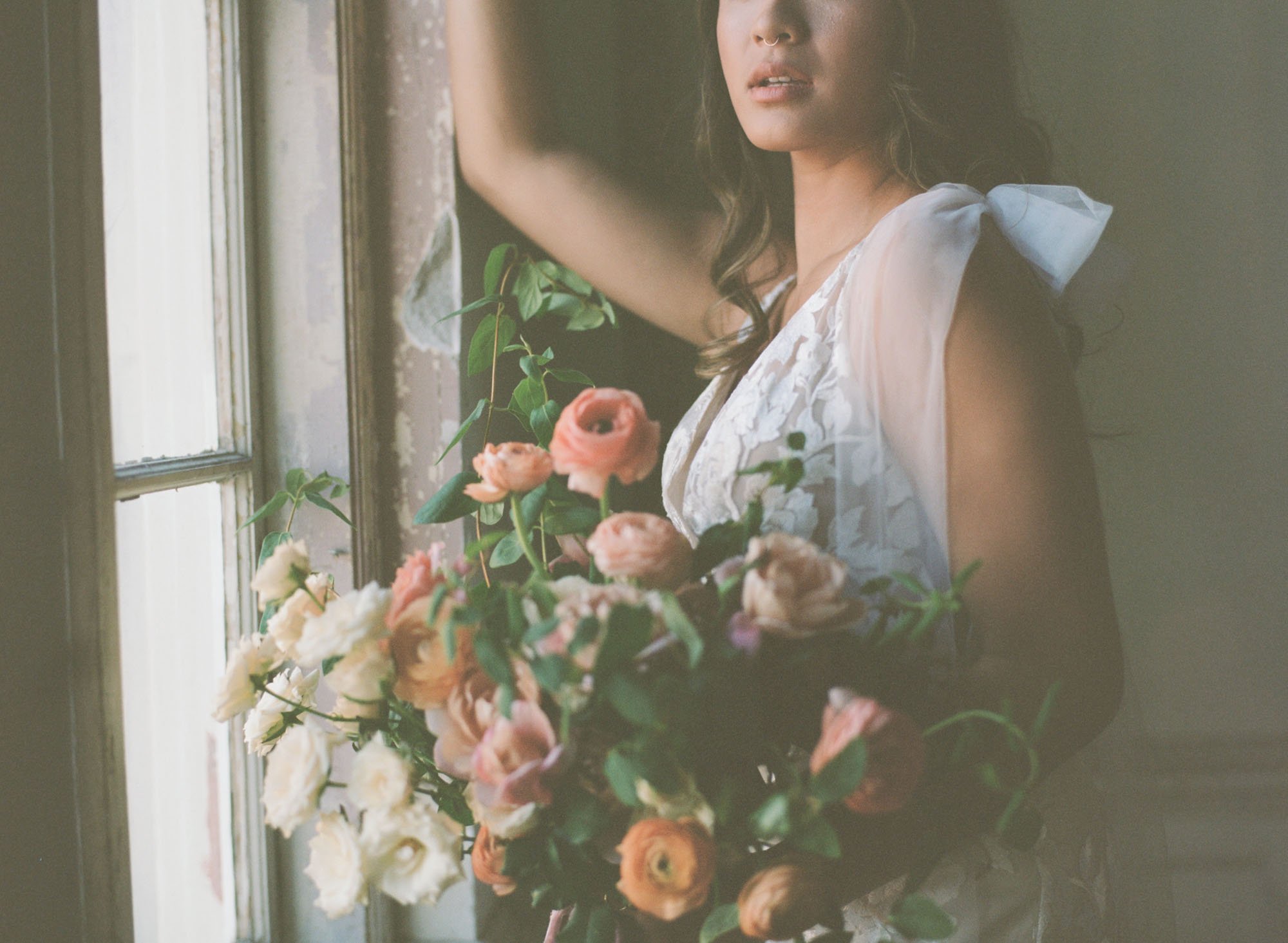  A bride wearing Made With Love Elsie and holding a large floral bridal bouquet consisting of pink ranunculus, white roses, and wild greenery. She is standing in front of an old window with one arm perched up on the window frame.  