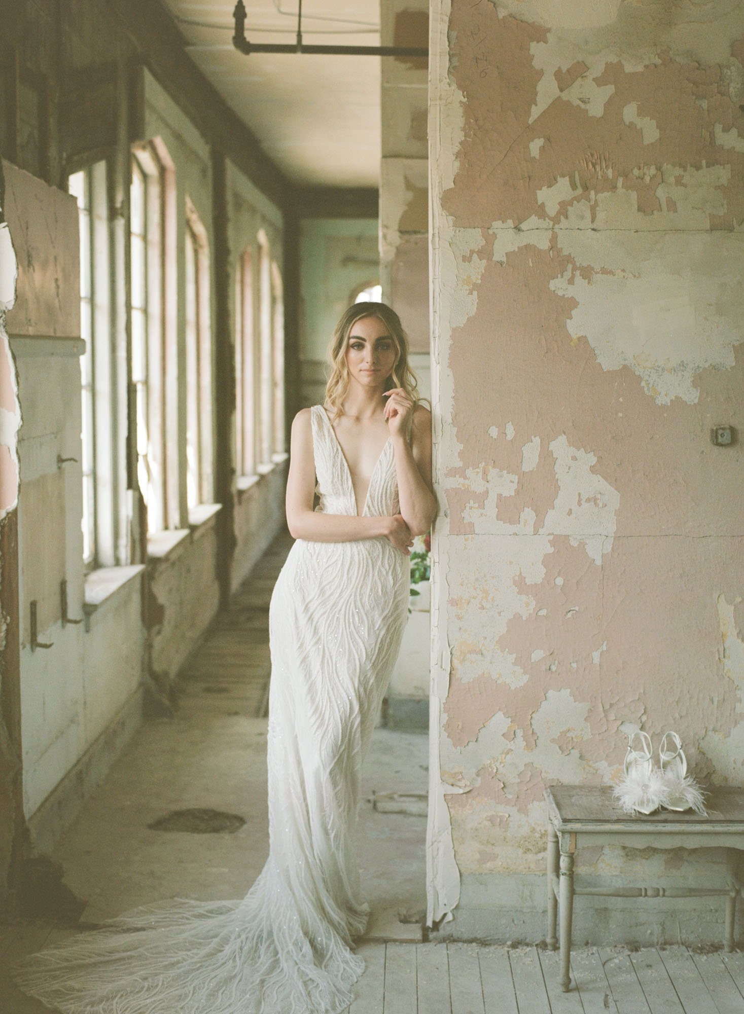  A bride wearing Made With Love Ryder wedding dress, which is a deep v fully. beaded fitted gown with a soft beaded train. She is standing in an old hotel and leaving against a wall in a hallway. Her white feather bridal shoes are sitting beside her.