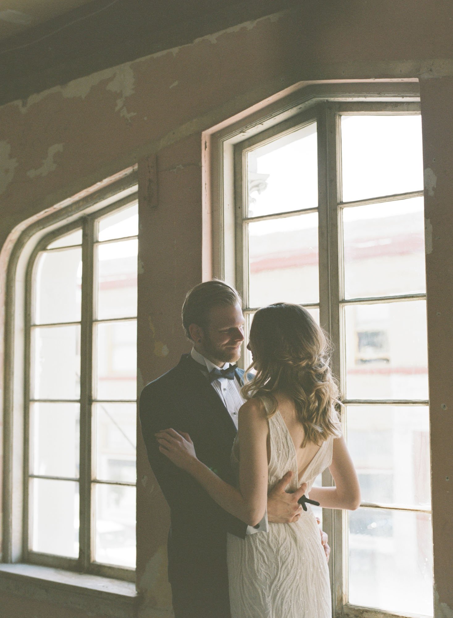  Photograph of a bride and groom embracing standing in front of a large bay window. The bride is wearing Made With Love Ryder and the groom is wearing a black Tuxedo.  