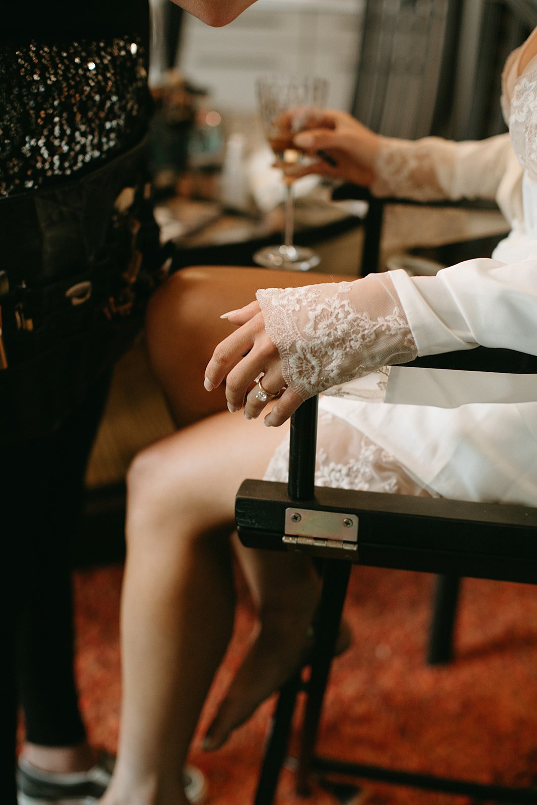  A bride getting ready for her wedding day wearing a white silk bridal robe with lace sleeves. She has a solitaire diamond engagement ring on her finger and a glass of champagne in her hand. 