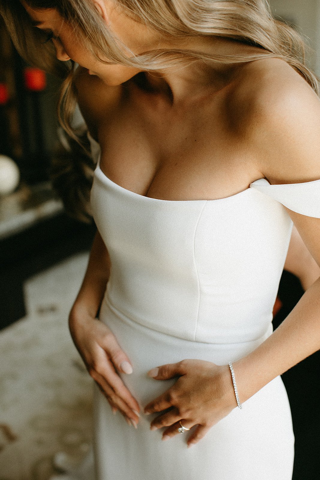  A bride wearing Alyssa Kristin Camila wedding dress, which is a fitted natural white silk crepe wedding dress with off the shoulder sleeves. She has long brown hair styled in soft curls and is wearing a diamond tennis bracelet. 