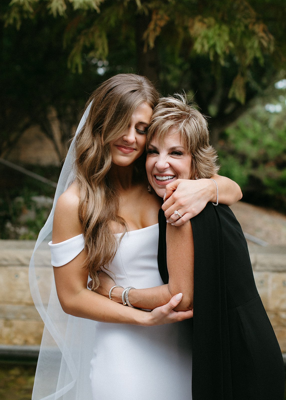  A bride wearing Camila wedding dress by Alyssa Kristin, hugging the mother of the bride who is wearing a chic sleeveless black dress. The bride is wearing the Julianne Veil by Sara Gabriel 