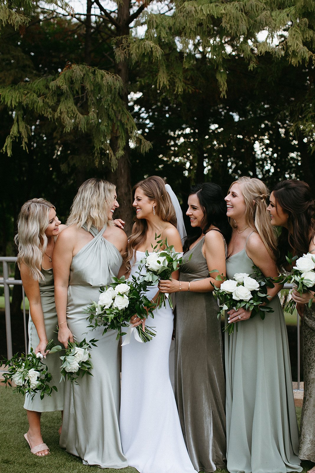  A bride wearing the Camila wedding dress by Alyssa Kristin, surrounded by bridesmaids wearing dresses in various shades of sage, mint, and pale green. They are all holding bouquets of large white roses and greenery smiling at each other. 