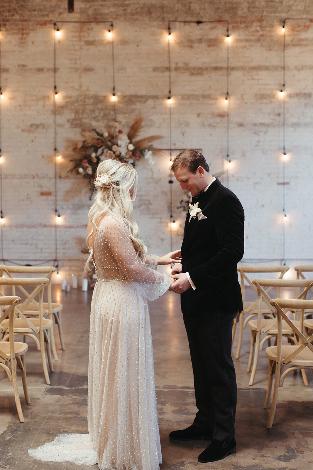  A bride wearing Lunella by Willowby by Watters having a first look with the groom on their wedding day. They are in a warehouse venue with light wash wood x-back chairs and hanging bistro lights in the background. 