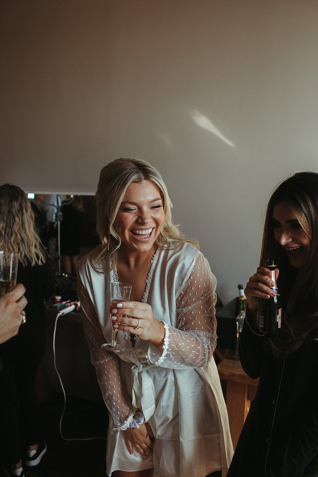  A bride laughing in a wedding robe drinking champagne with their bridesmaids. 