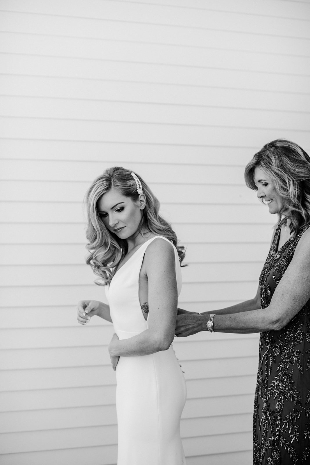  The mother of the bride zipping the bride into their Alyssas Kristin Maven wedding dress on their big day. The bride has long blonde hair styled in soft bridal waves with a pearl hair clip. 