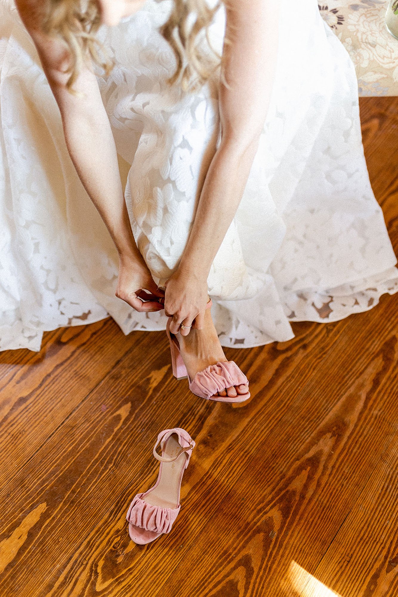  A bride wearing Truvelle Annie wedding dress bending over to fasten the ankle strap on a pair of pink suede ruched bridal shoes. 