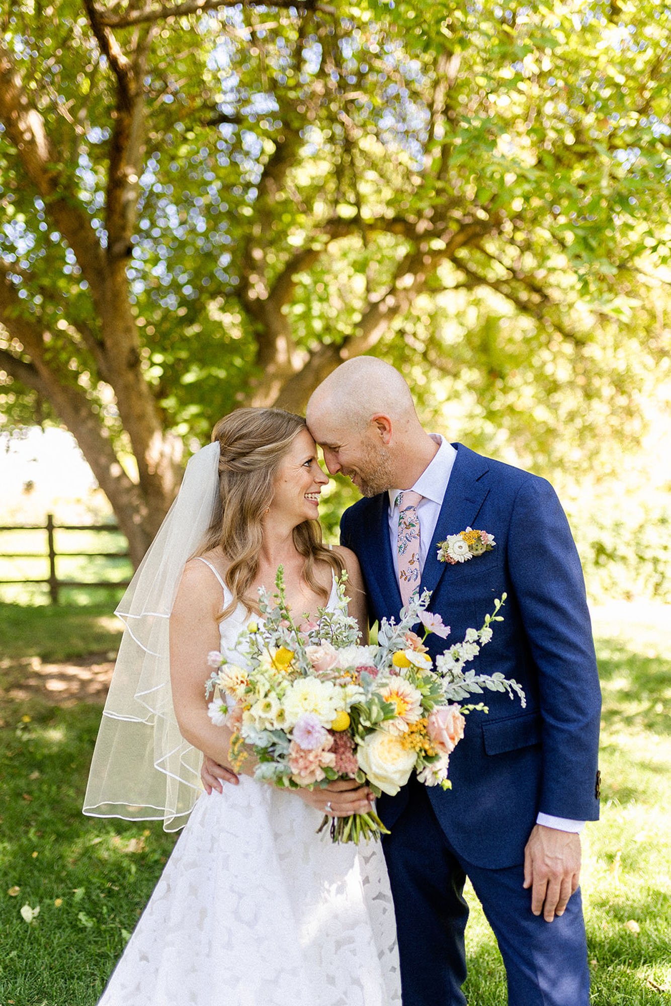  A bride and groom standing side by side facing each other and smiling. The bride is wearing the Annie wedding dress by Truvelle bridal and is holding a large springtime bridal bouquet. 