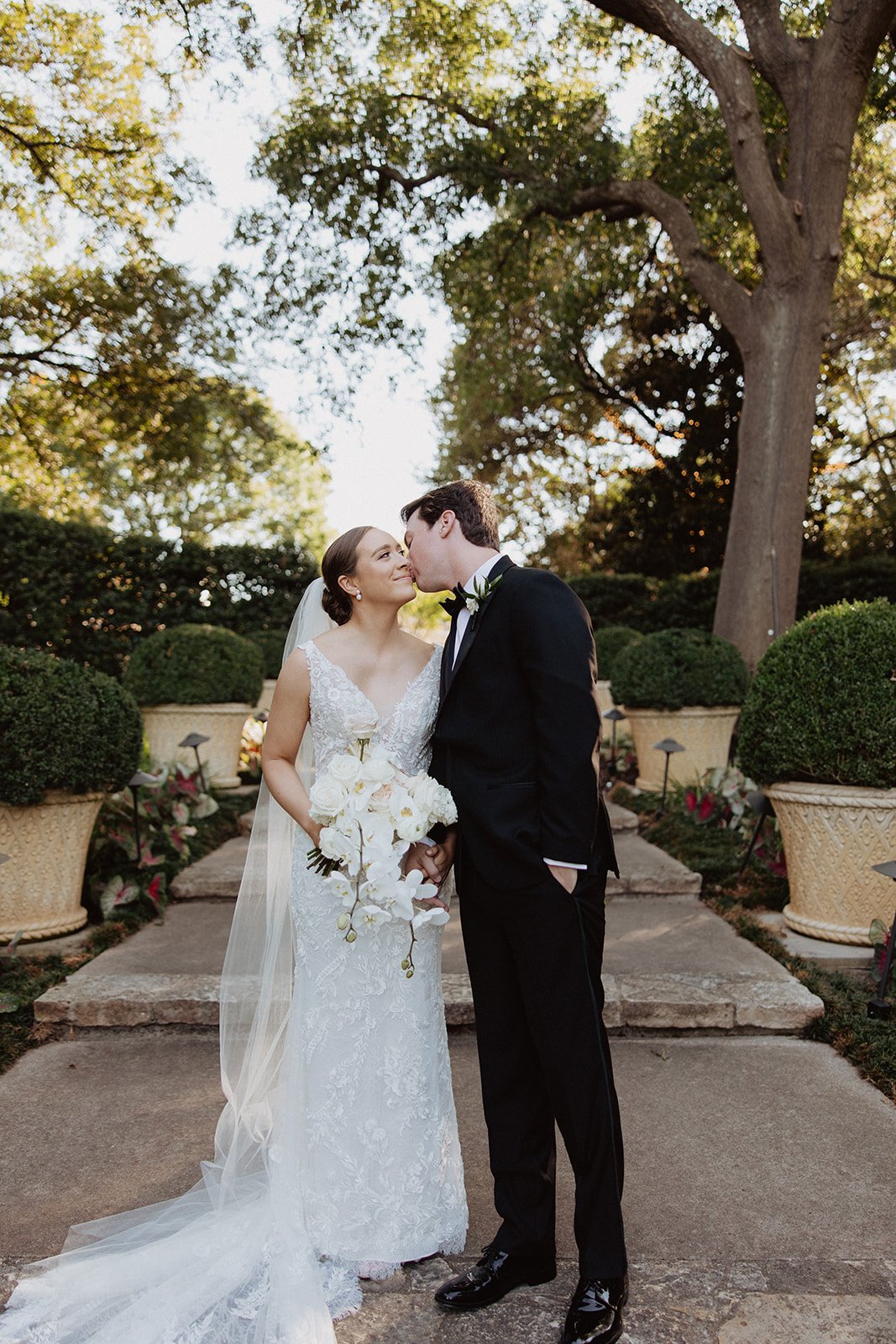  A bride wearing ‘lucia’ by Wtoo while the groom kisses her on the cheek at the Dallas Arboretum and Botanical Garden  