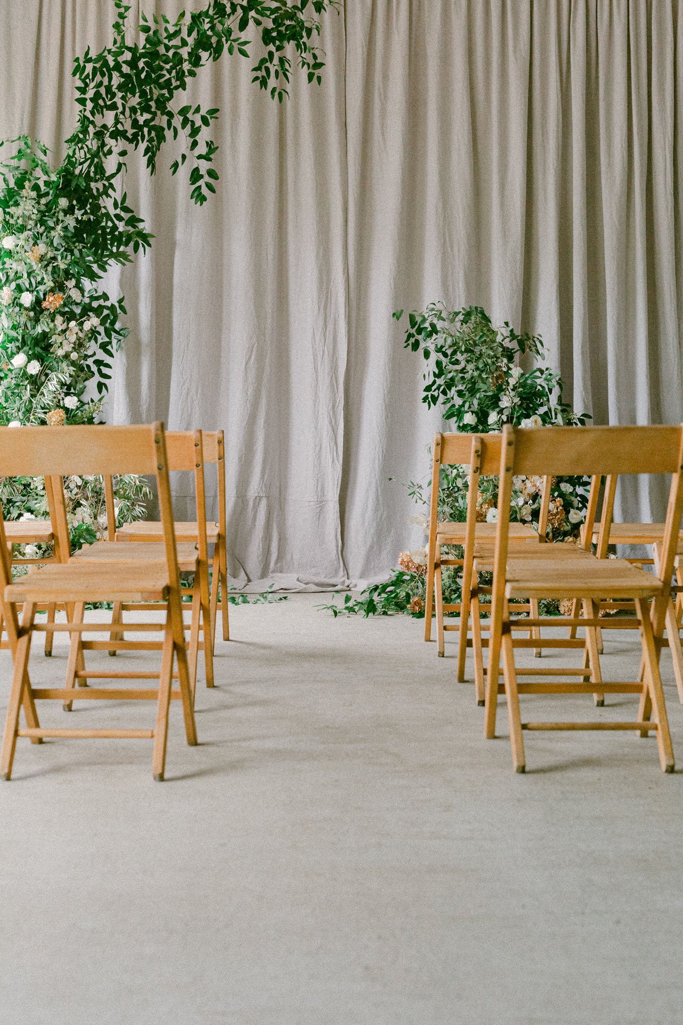 A lovely and intimate ceremony setup with worn wooden folding chairs, draped linen backdrop, and wild greenery arbor installation 