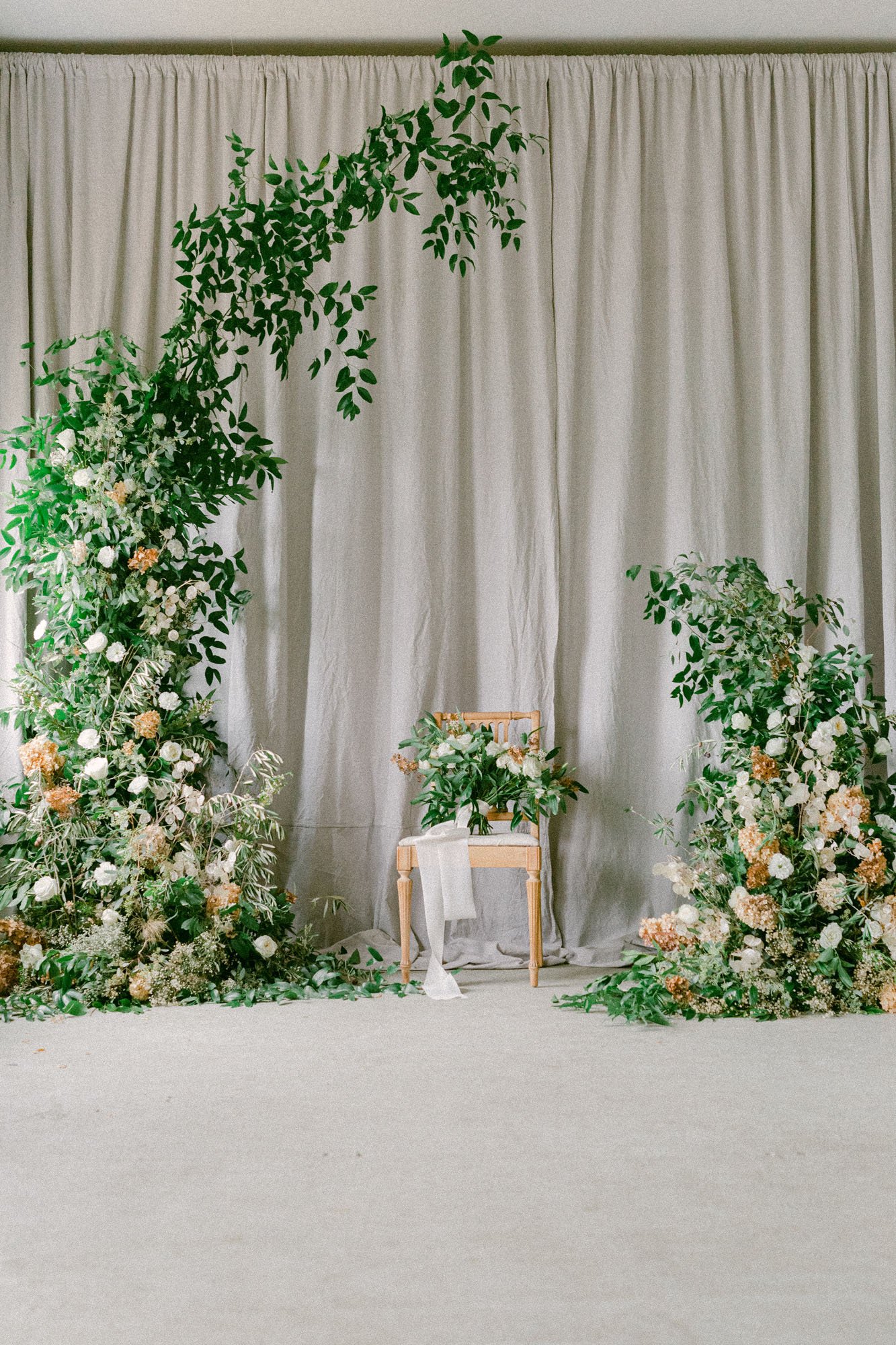  a wild and organic wedding arbor installation with lovely greenery, dried florals, and large white blooms in front of a grey linen backdrop 
