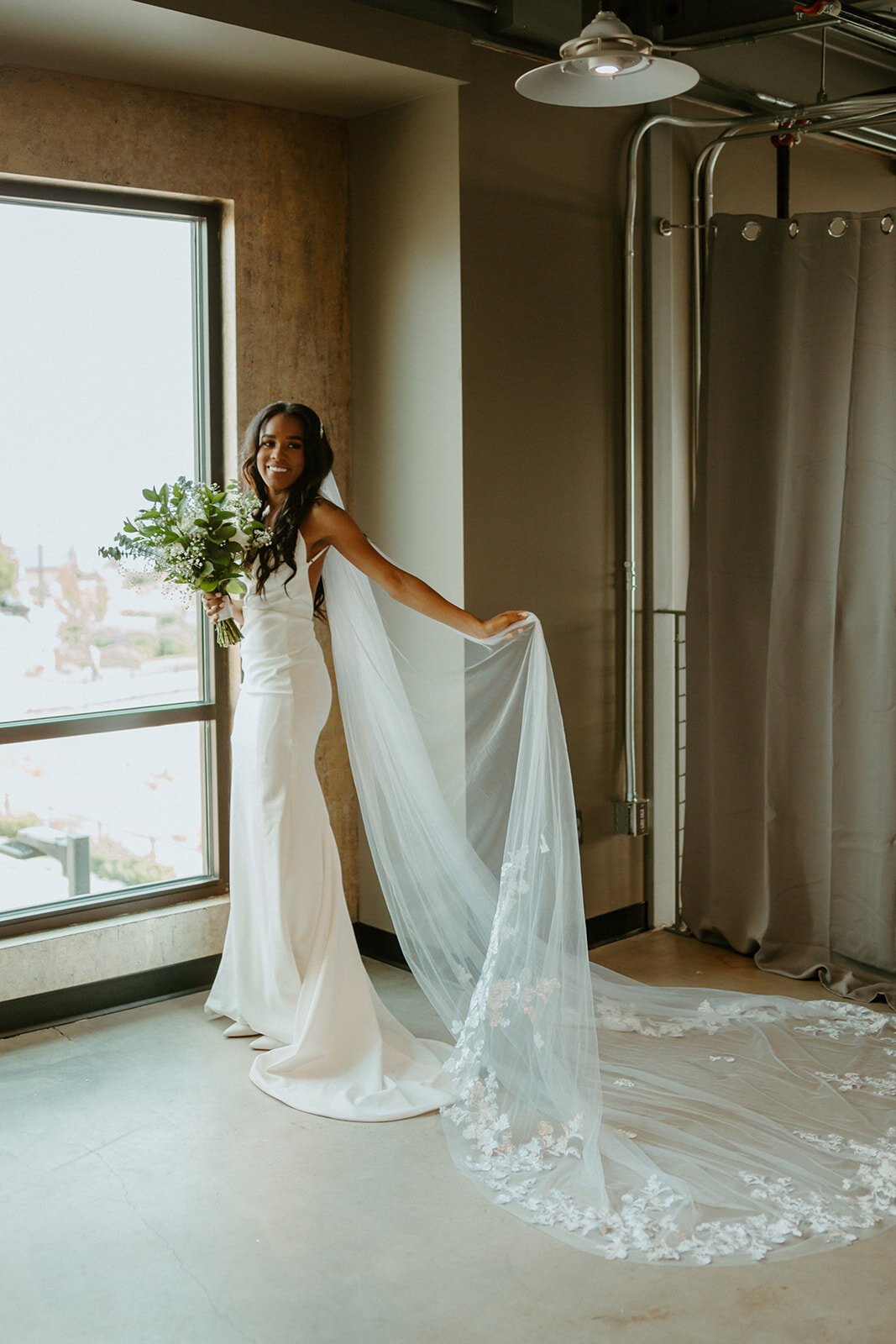  A bride wearing TENNYSON by The Label wedding dress, standing in front of a large window smiling and wearing a long veil with lace trim 