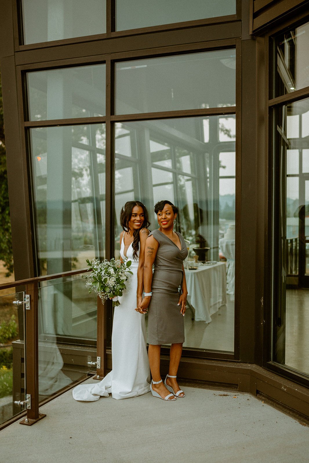  A bride and mother of the bride standing side by side on a balcony smiling. The bride is wearing TENNYSON by The Label. 