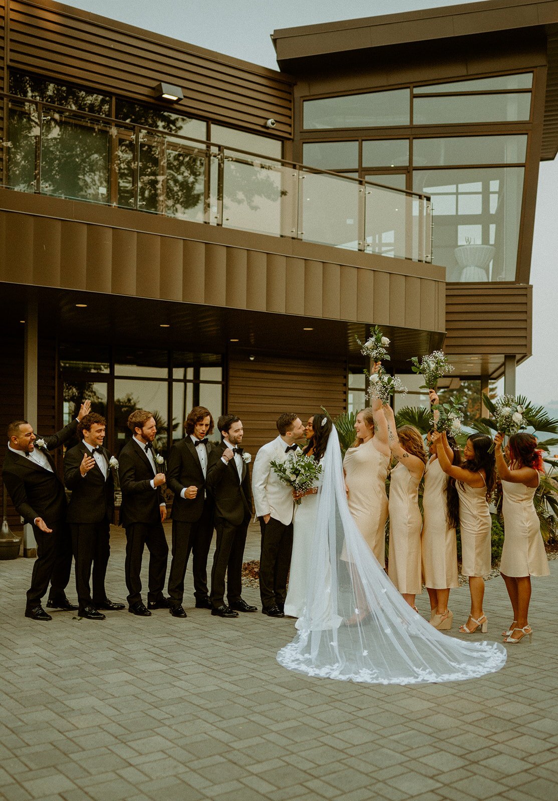  A bridal party standing on a waterfront patio. The bride is wearing TENNYSON by The Label, the groom is wearing a white tux, and the bridesmaids are wearing champagne silk dresses and the grooms are wearing black tuxedos.  