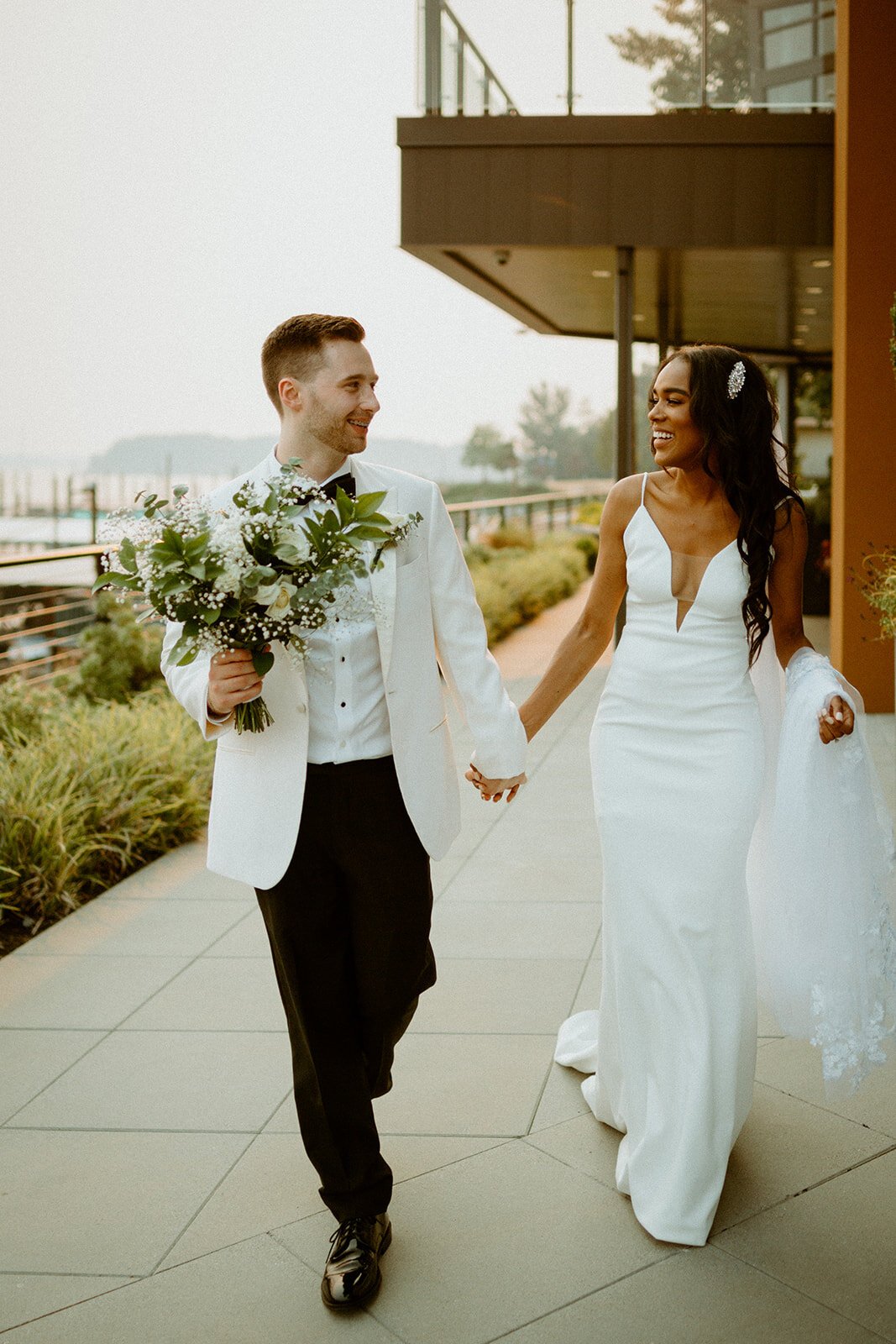  A bride wearing the deep-v fitted spaghetti strap wedding dress, TENNYSON, by The Label walking down the water front holding hands with the groom who is wearing a white tux and holding the bride’s bouquet 