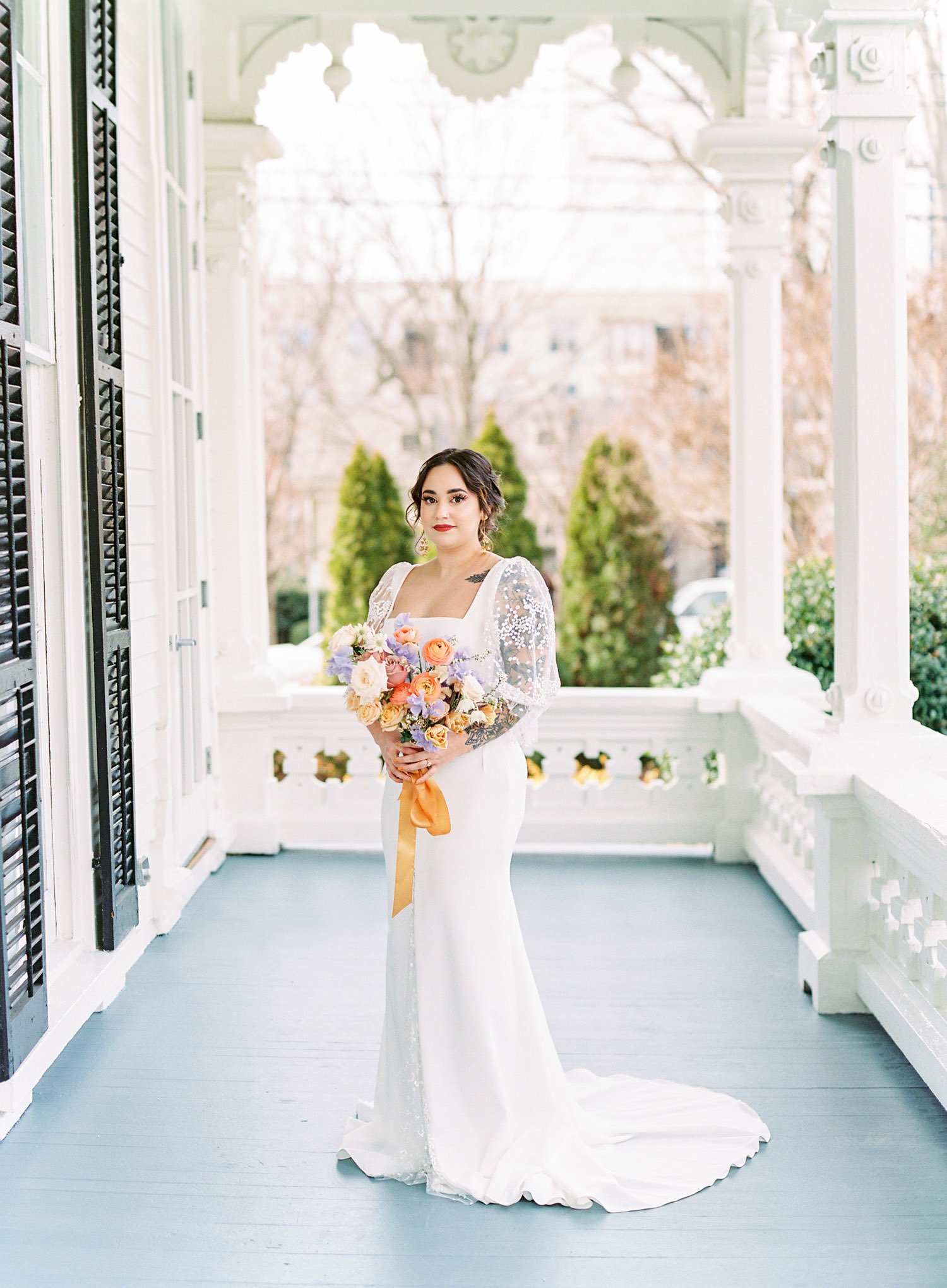  a bride standing on the porch of merrimon-wynne house in raleigh north carolina wearing the romantic florance wedding dress by laudae, a square neck long sheer puff sleeve bridal gown with a center slit and inset lace and low back 
