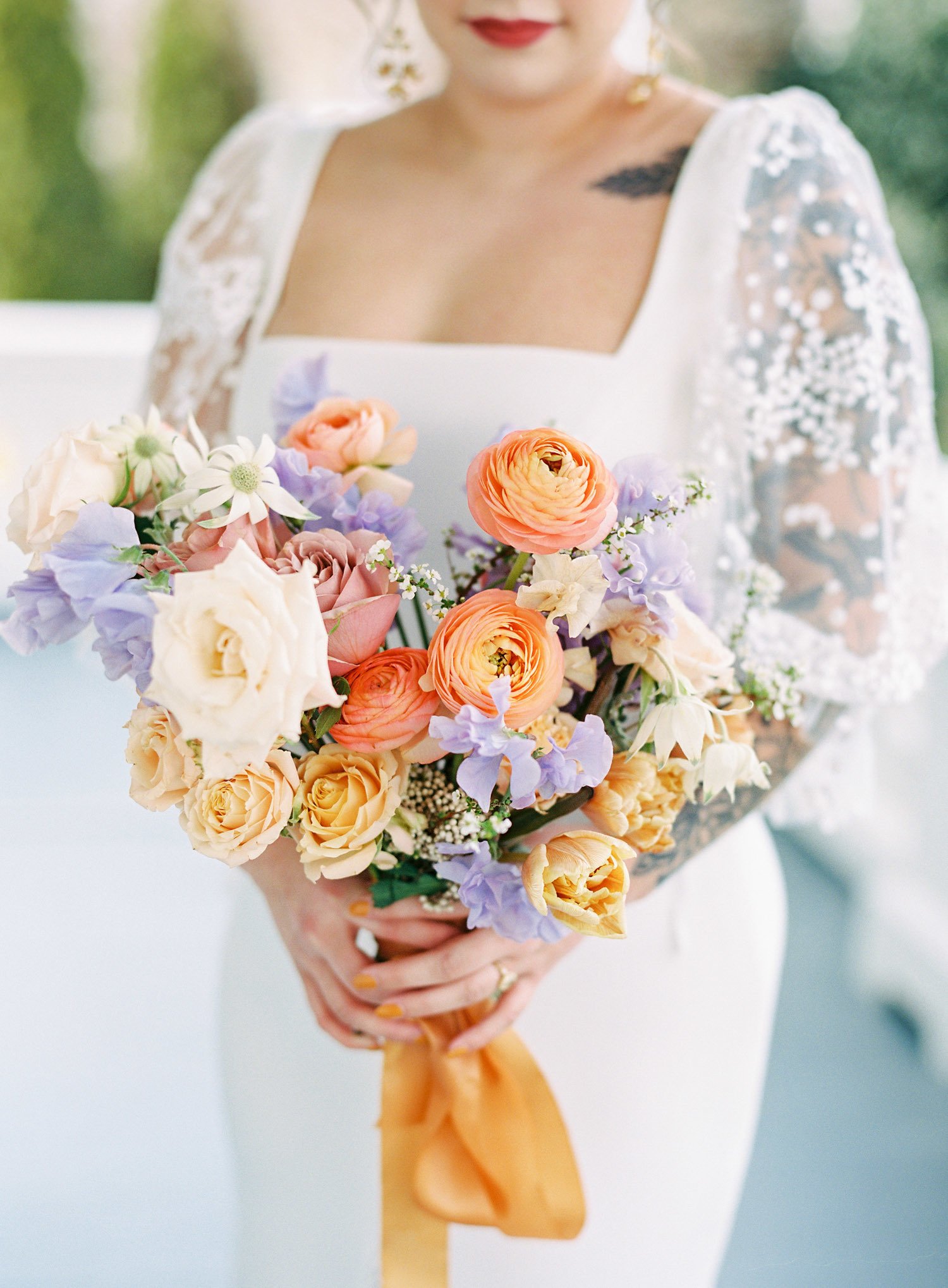  a bride holding a bouquet of peach, cream, orange, purple and white with an orange silk ribbon standing on the porch of the merrimon-wynne house in raleigh north carolina 