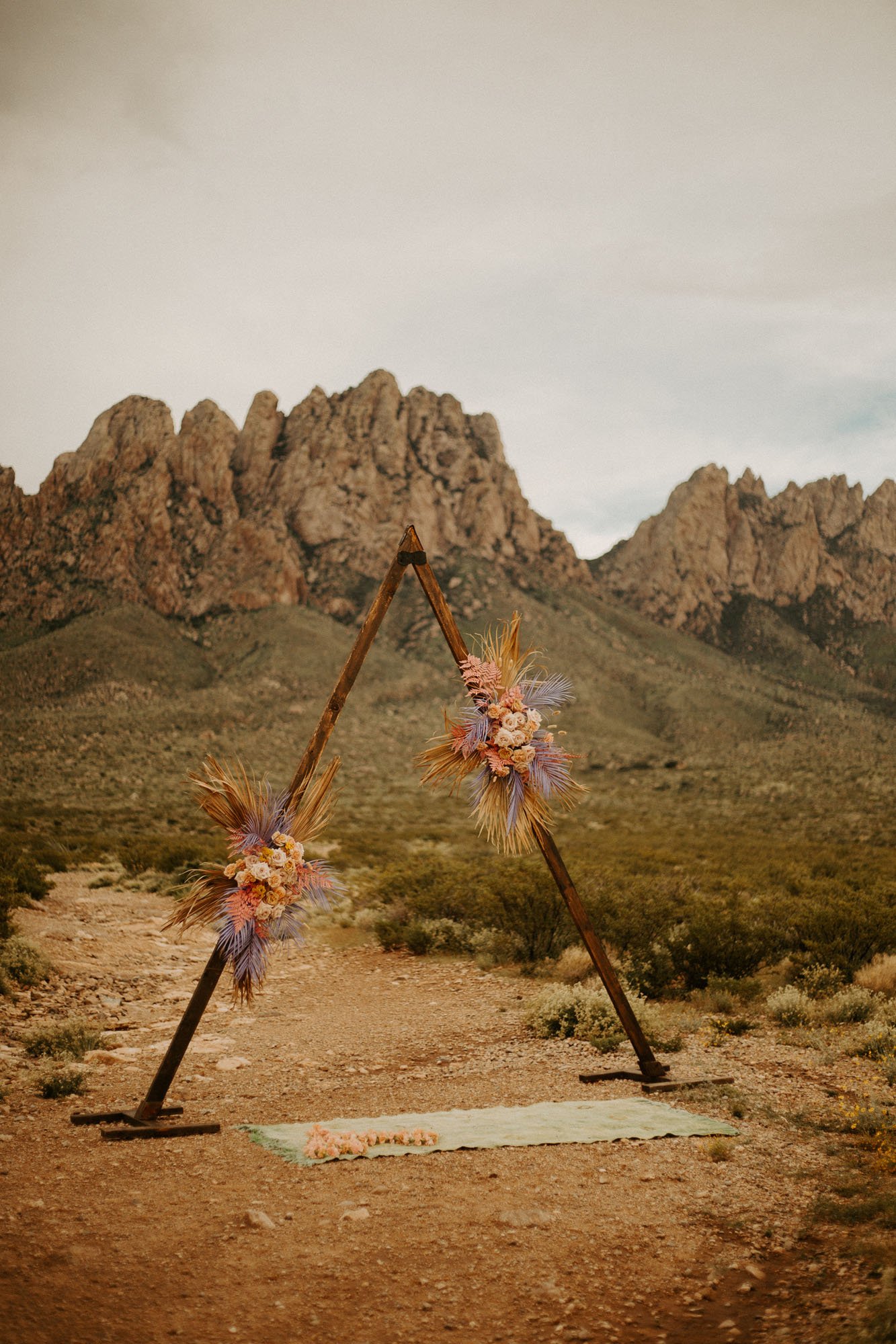  a fun triangle wedding arbor for a new mexico boho elopement with dried florals and peach, cream, and purple greenery in front of a mountain backdrop 