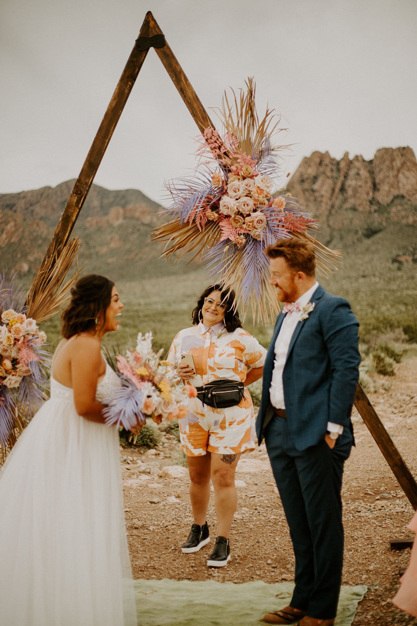  a bride and groom laughing while exchanging vows under a triangle wedding arbor for their desert elopement in new mexico. the bride is wearing thistle by willowby by watters and the groom is wearing a blue plaid suit. 