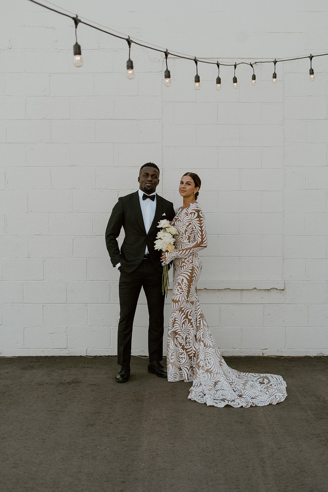  a bride and groom standing against a white brick wall under romantic bistro lights for their first look. the bride is wearing a lovely bold lace wedding dress by rue de seine bridal. 