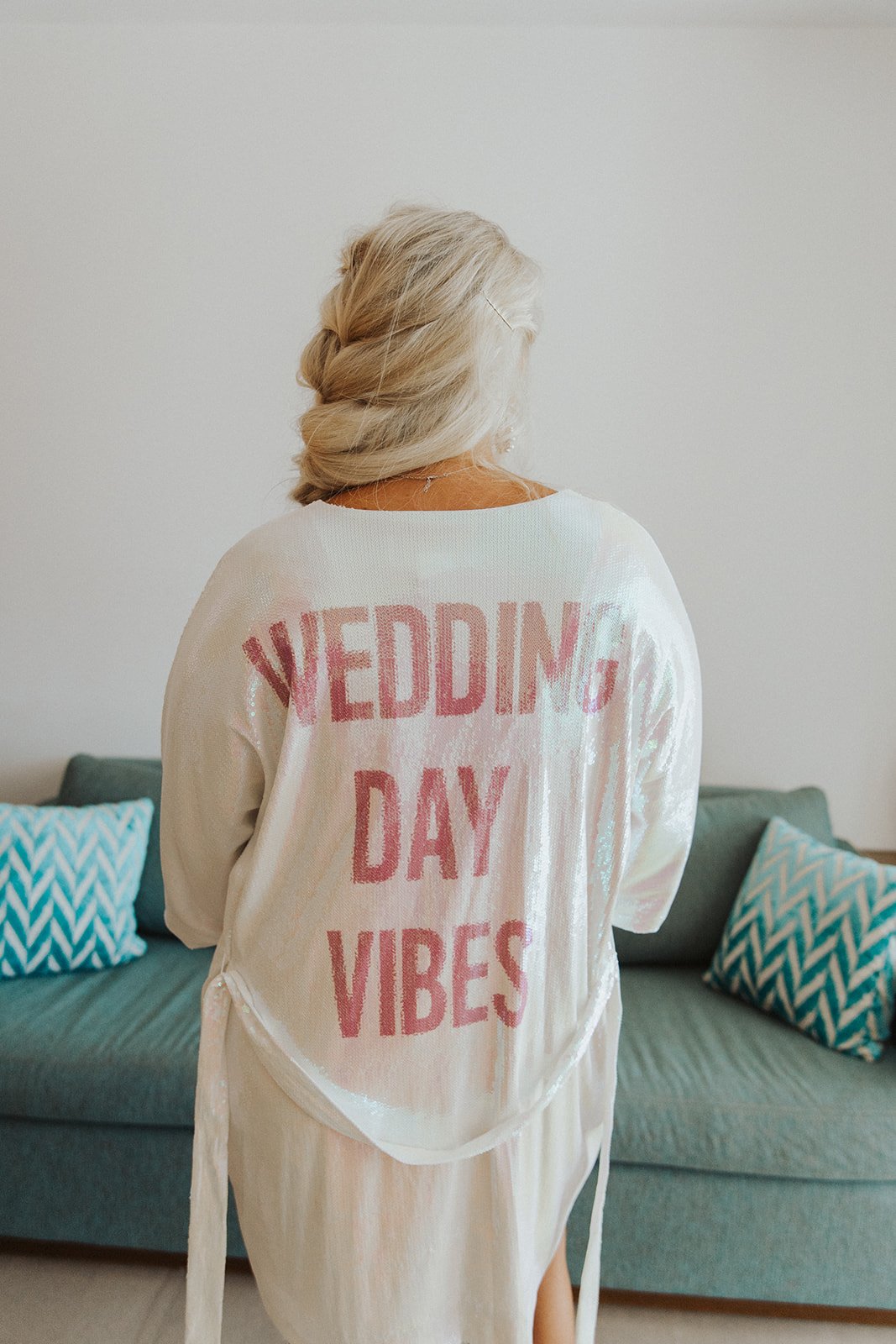  A bride wearing a white robe that says “wedding day vibes” in pink sequins on the back. 