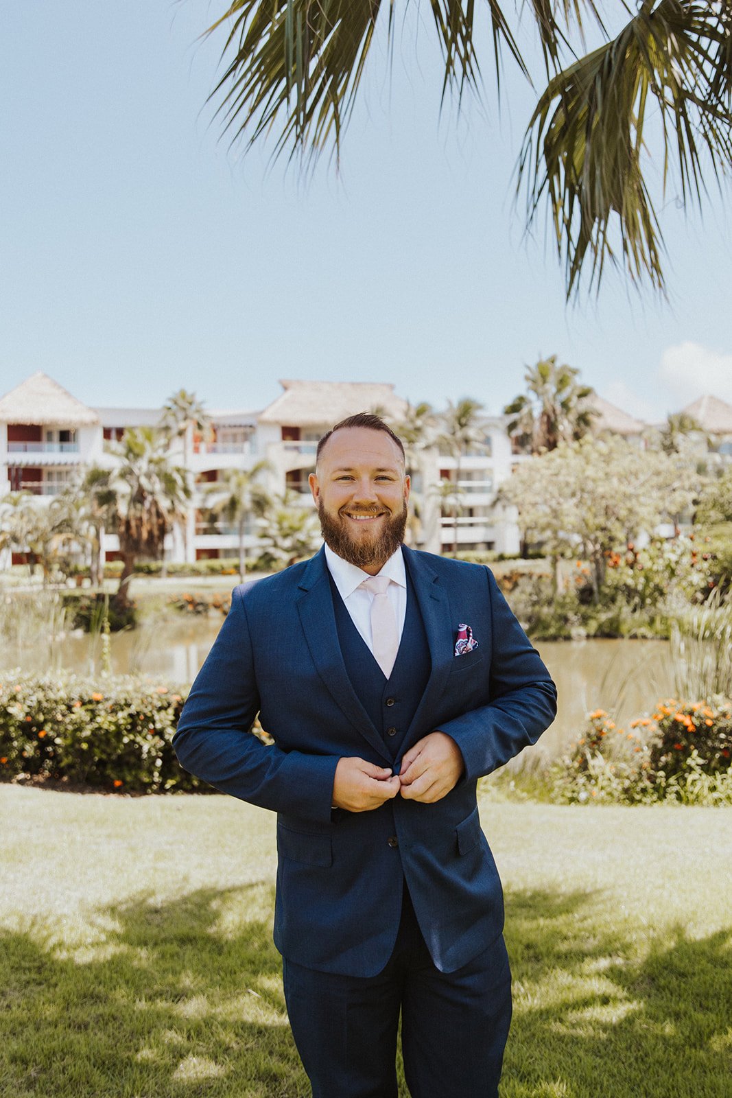  A groom standing in front of a tropical water way wearing a blue suit and smiling 