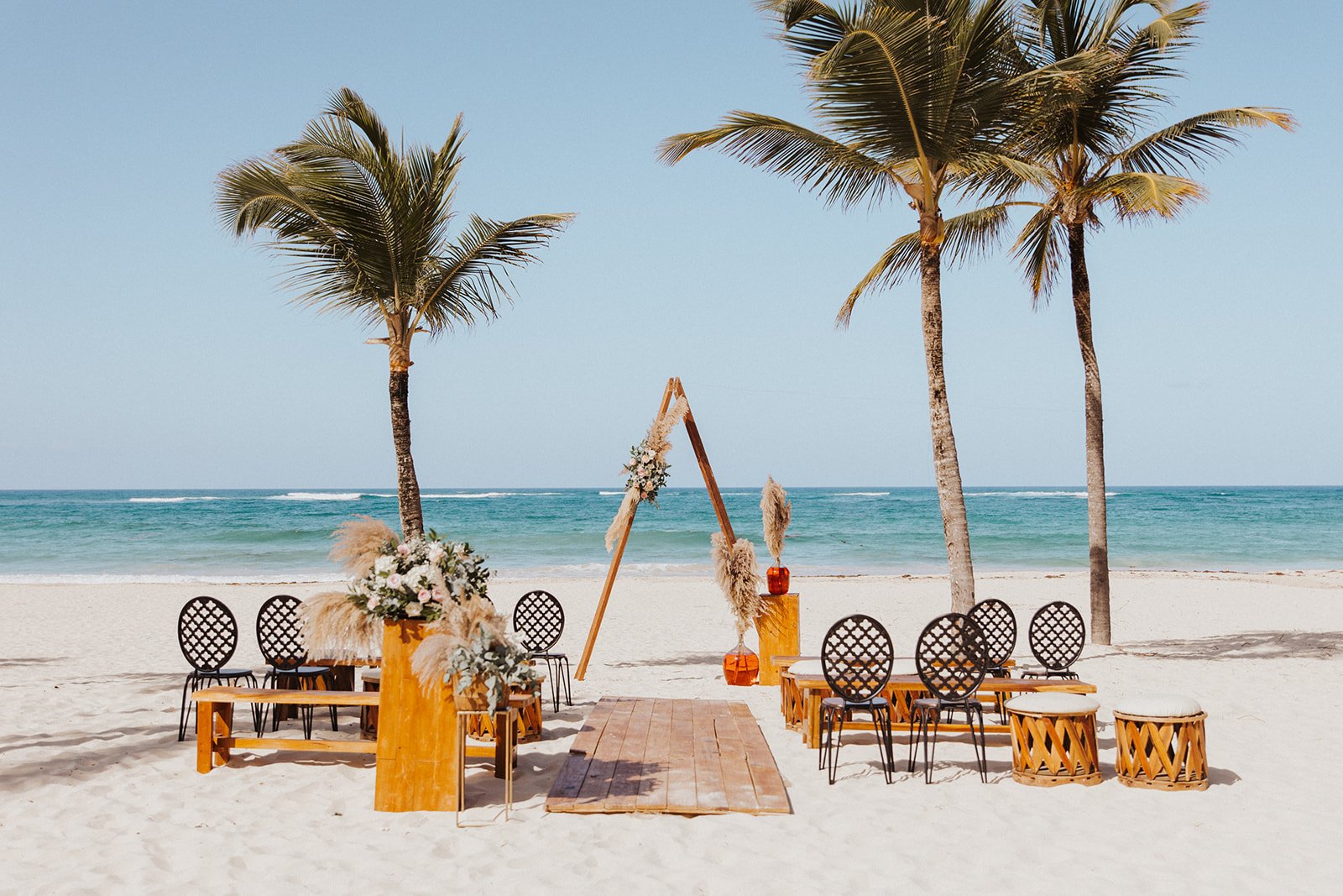  A beach destination wedding ceremony setup with a lovely boho arbor with pampas grass and florals under palm trees with the ocean in the background 