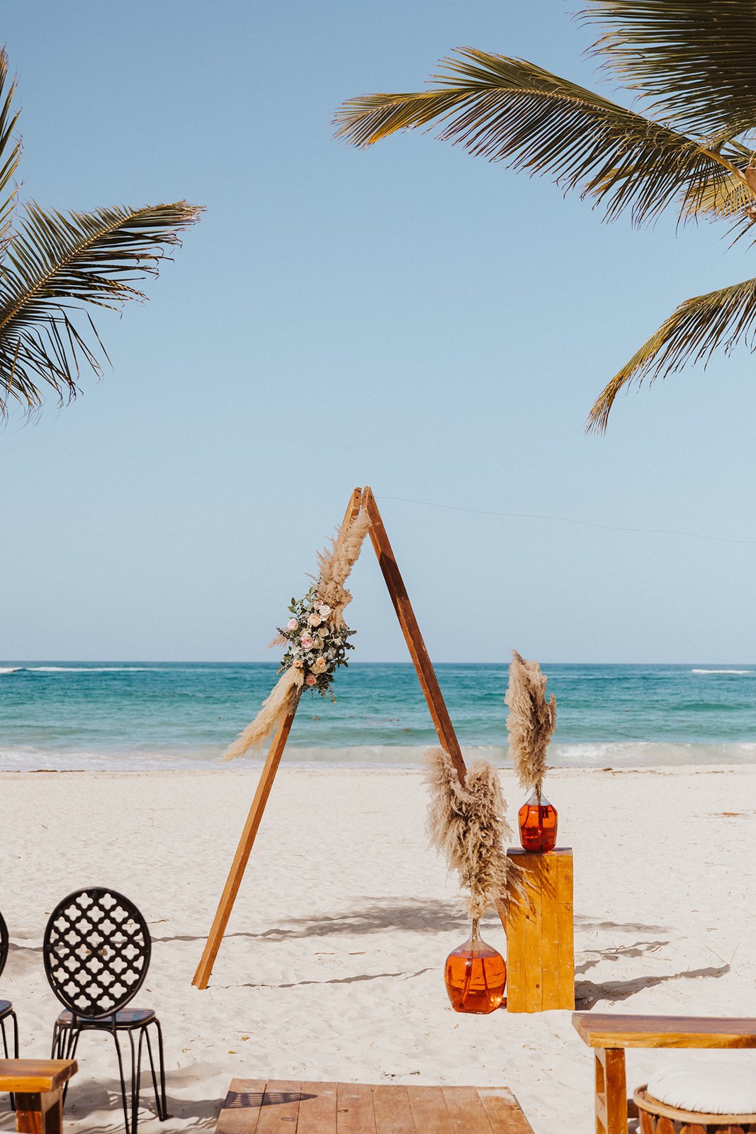  A fun boho ceremony arbor on the beach styled with pampas grass and florals. The ocean and palm trees are in the background. 