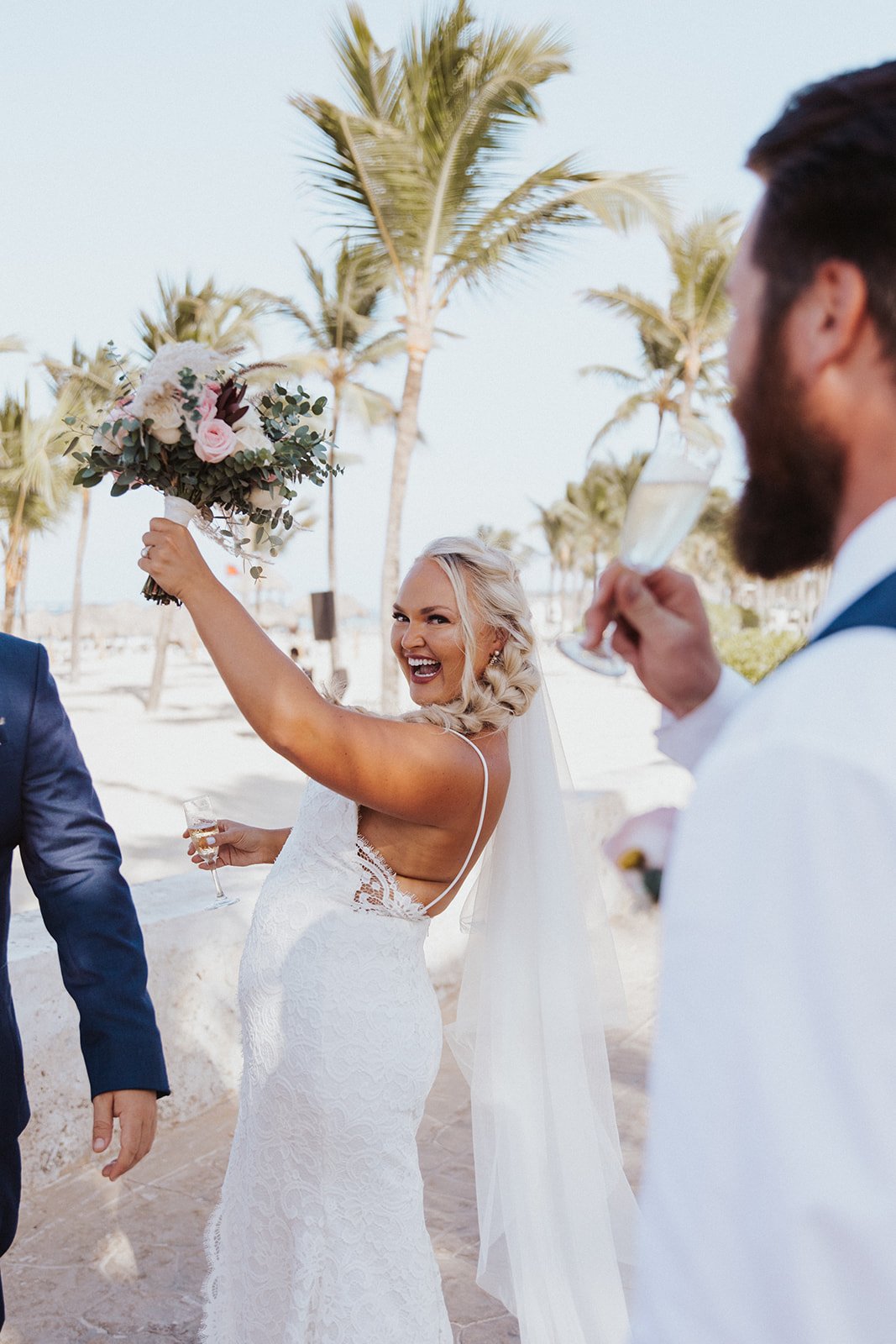  A bride smiling wearing Anais Anette ‘Emmanuelle wedding dress and holding up a lovely bridal bouquet after saying “I do” on the beach 