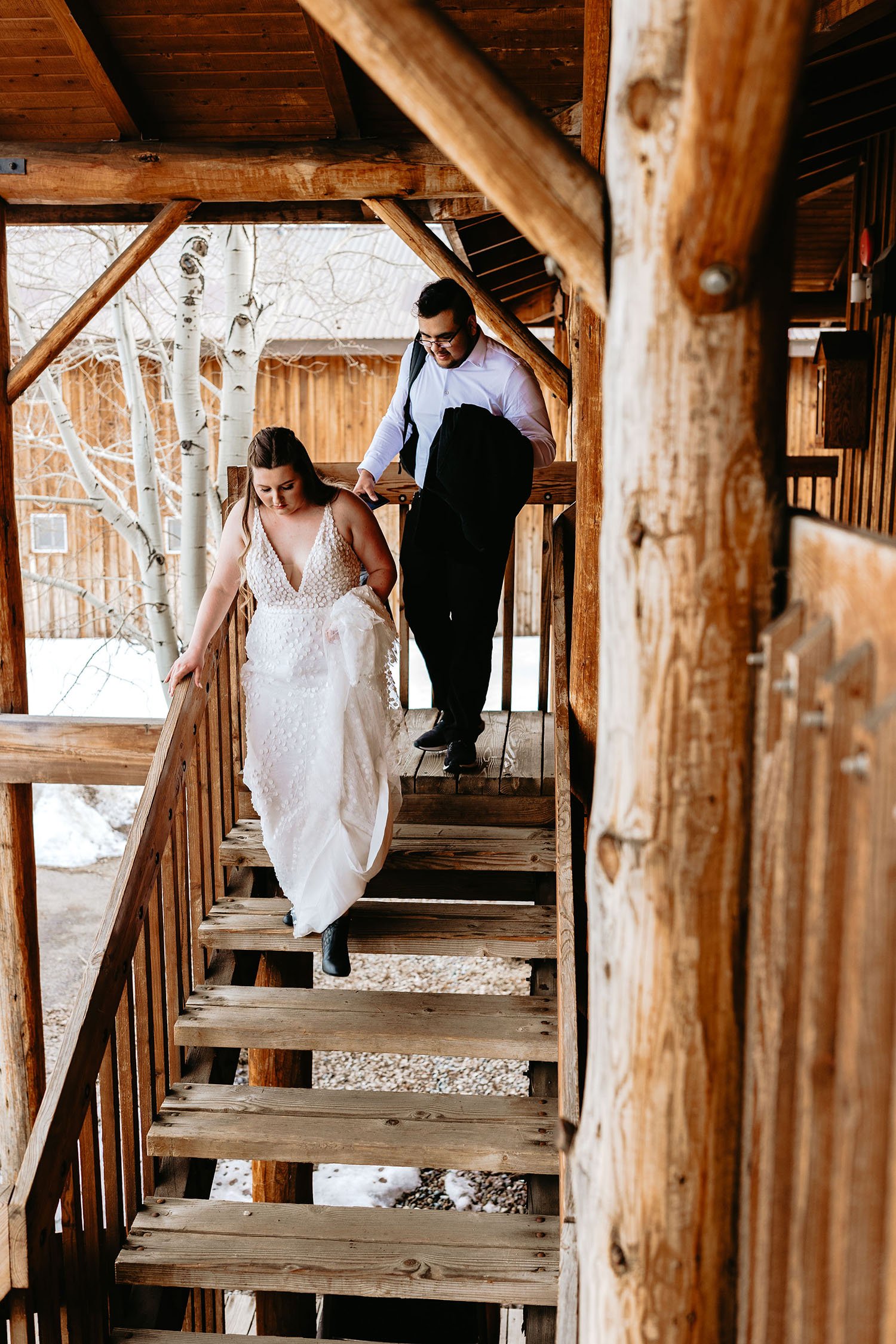  A bride wearing Made With Love ‘Louie’ wedding dress for a snowy Idaho elopement, walking down the steps of a rustic cabin with the groom on the way to exchange their vows 
