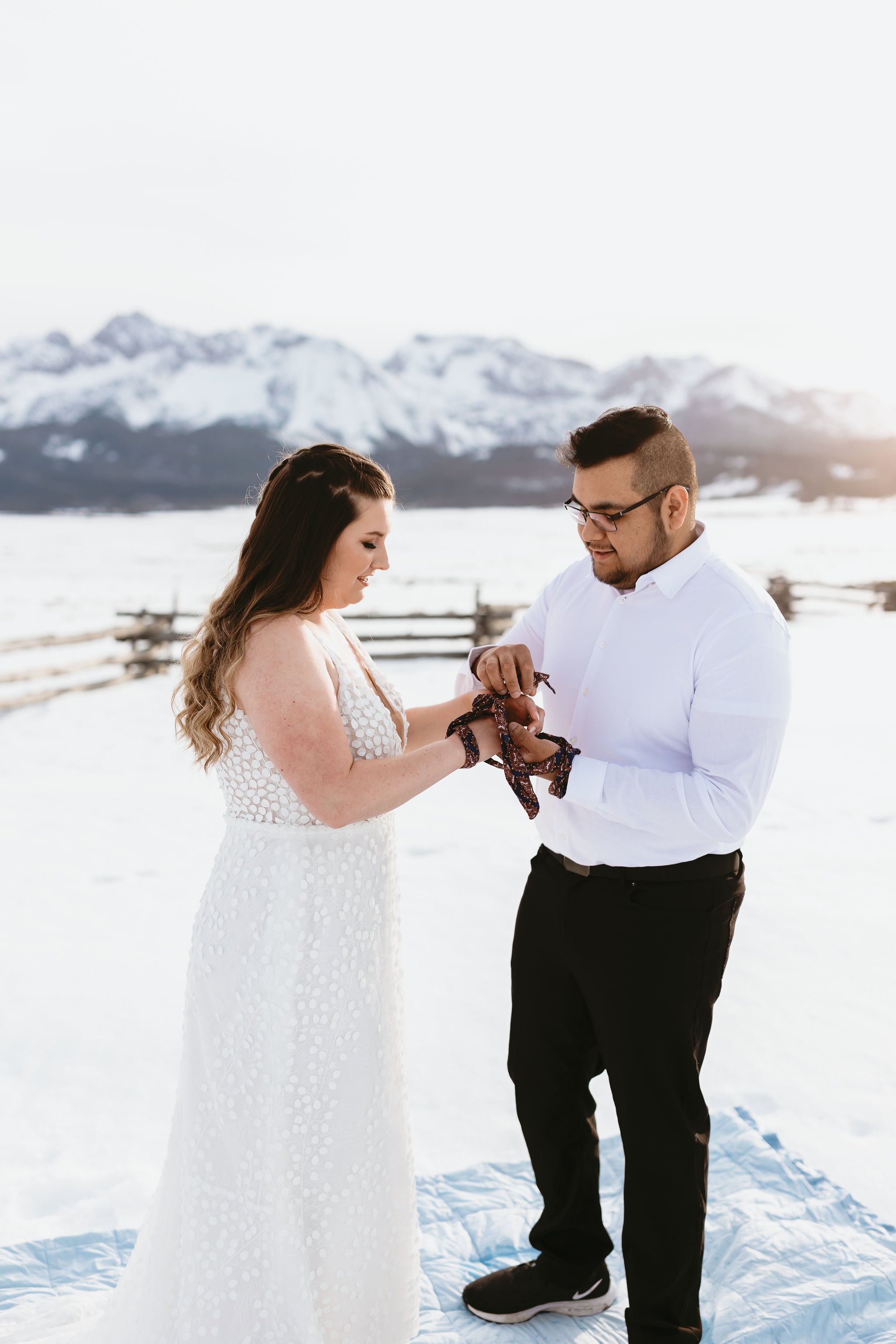  A bride and groom performing a handfasting ceremony for their snowy Idaho elopement in the mountains. The bride is wearing the romantic ‘Louie’ wedding dress by Made With Love bridal. 