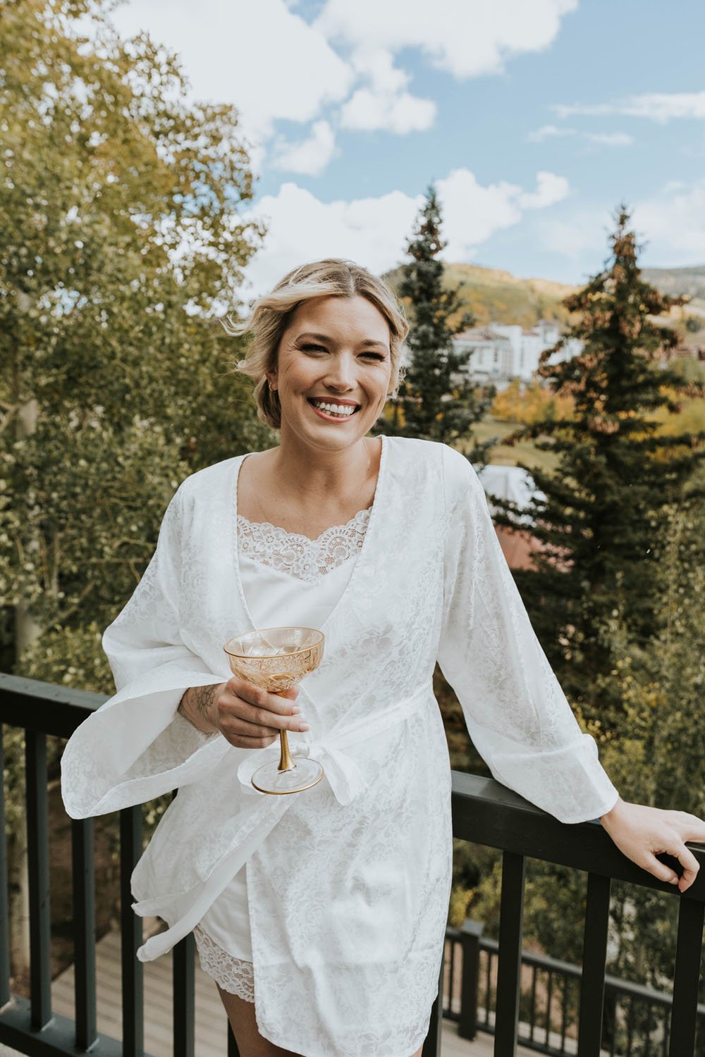  a bride wearing a white lace and silk bridal robe smiling on a balcony overlooking the mountains in telluride colorado while getting ready for her wedding. 