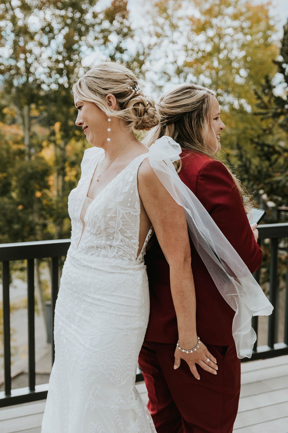  two brides sharing an emotional first look before their wedding ceremony. one bride is wearing the lovely ‘selene’ wedding dress by rish, and the other bride is wearing a burgundy suit. they are standing on a cabin balcony in telluride, colorado 