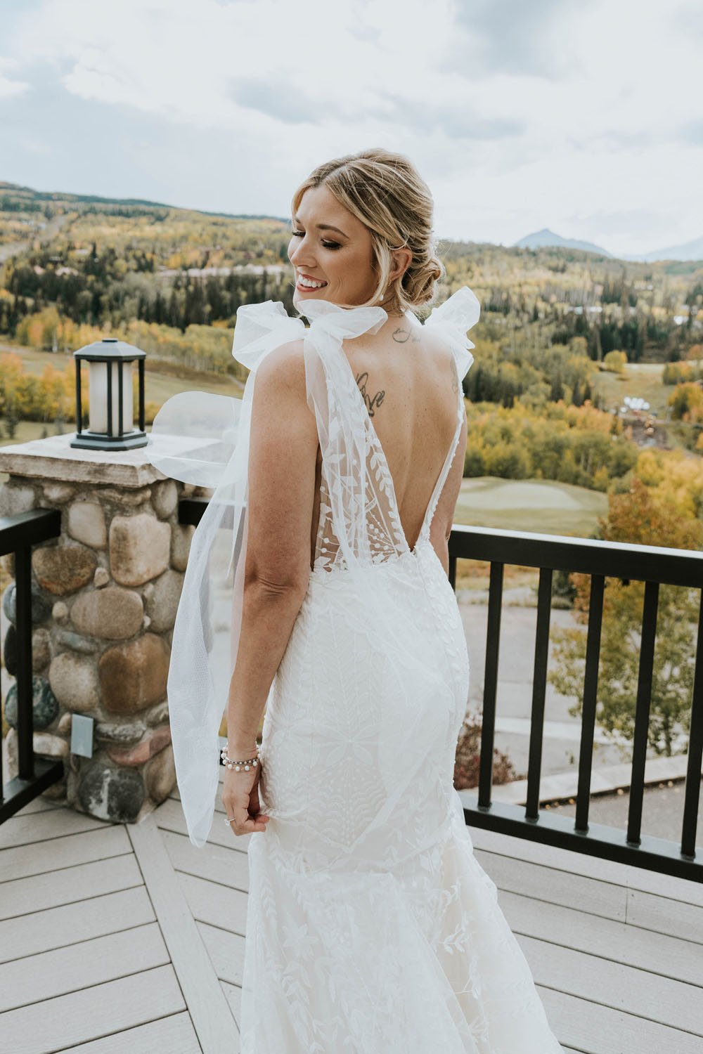  a bride looking over her shoulder smiling while wearing a low v-back wedding dress with illusion lace and shoulder bows by made with love 