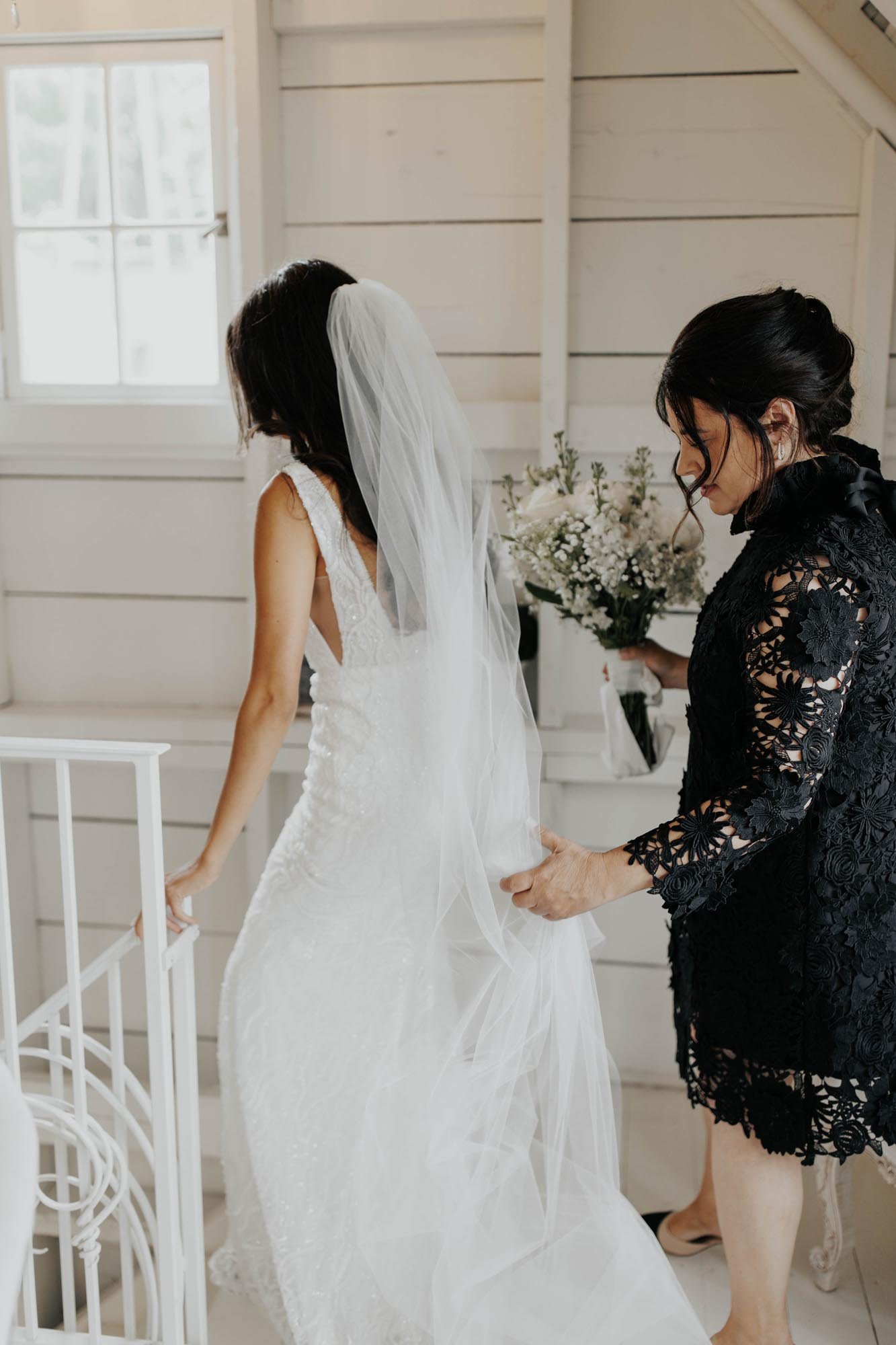  A bride walking down the stairs in her wedding gown and veil with help from her mother carrying her bridal bouquet.  