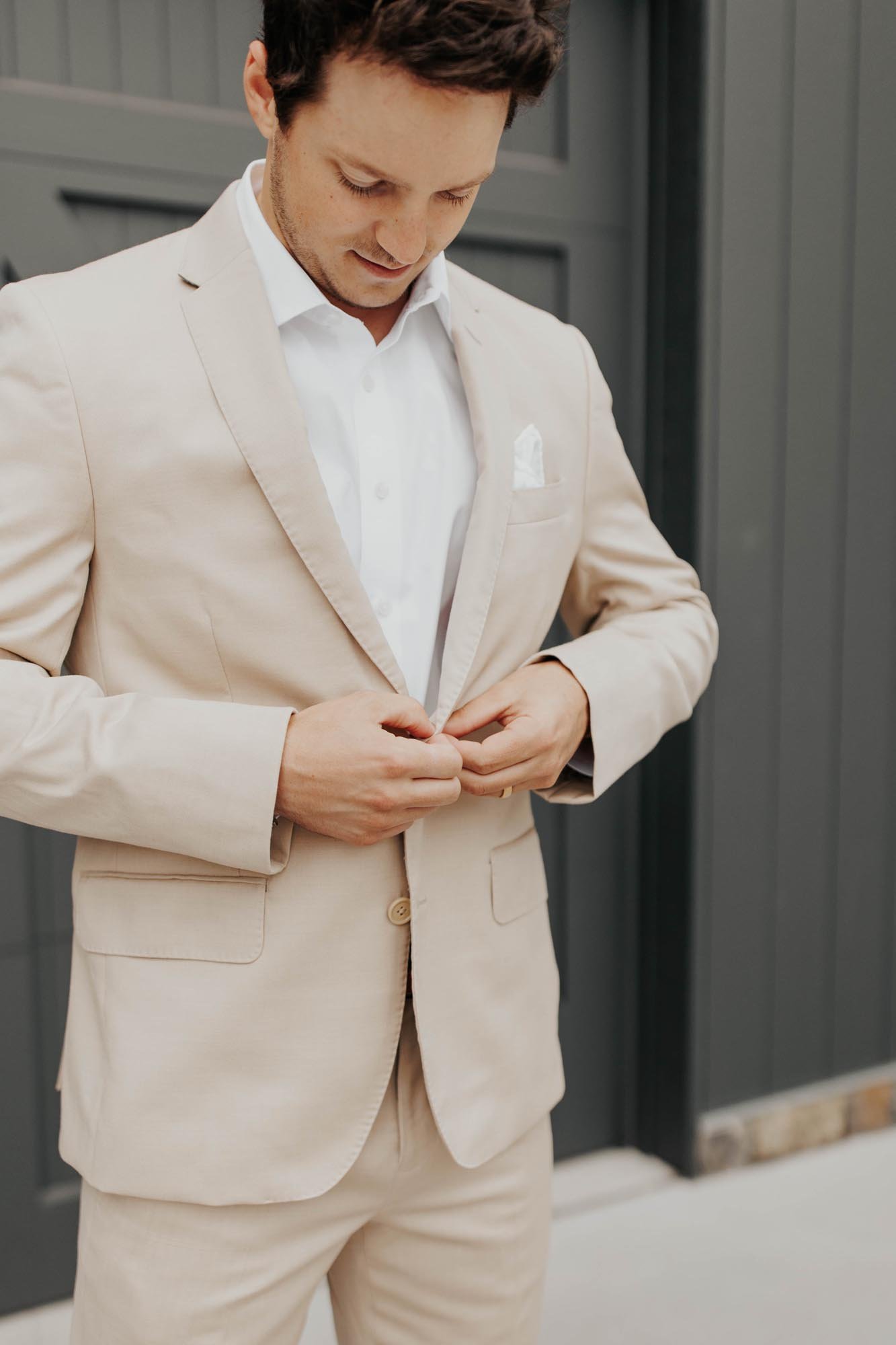  A groom wearing a tan suit buttoning his jacket standing in front of a black painted barn while waiting for the first look with his bride.  