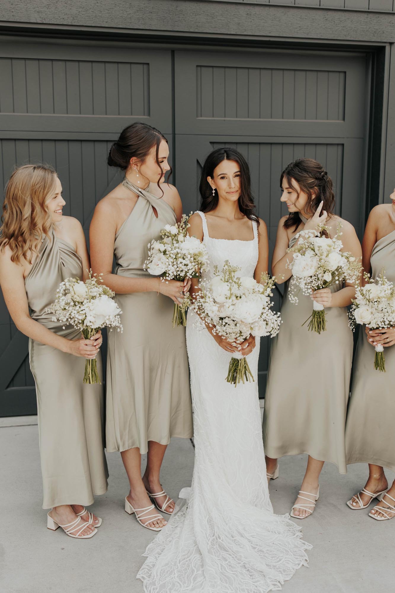  A bridal party wearing champagne satin halter nek bridesmaids dresses surrounding the bride wearing a square neck fitted beaded wedding dress. They are all holding bouquets with white roses, peonies, and babies breath and standing in front of a blac