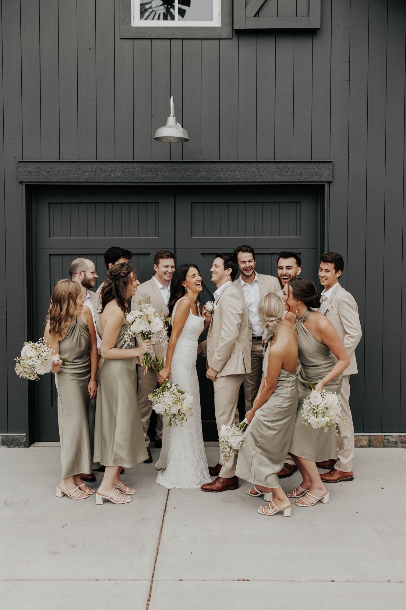  A wedding party laughing together before the ceremony. The bride is wearing a white beaded wedding dress, bridesmaids are in champagne satin midi dresses, and the groom and groomsmen are in tan suits.  