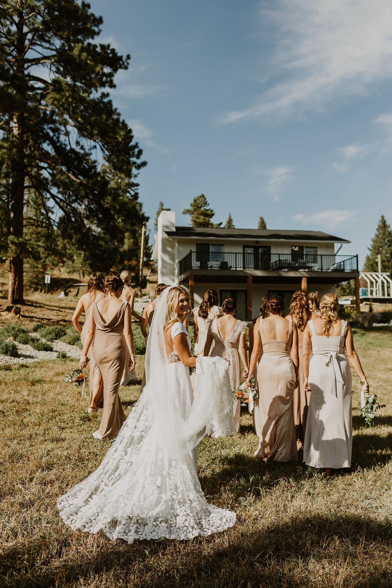  a bride wearing a white lace wedding dress and long lace veil is walking with her bridesmaids towards their cabin wedding venue 