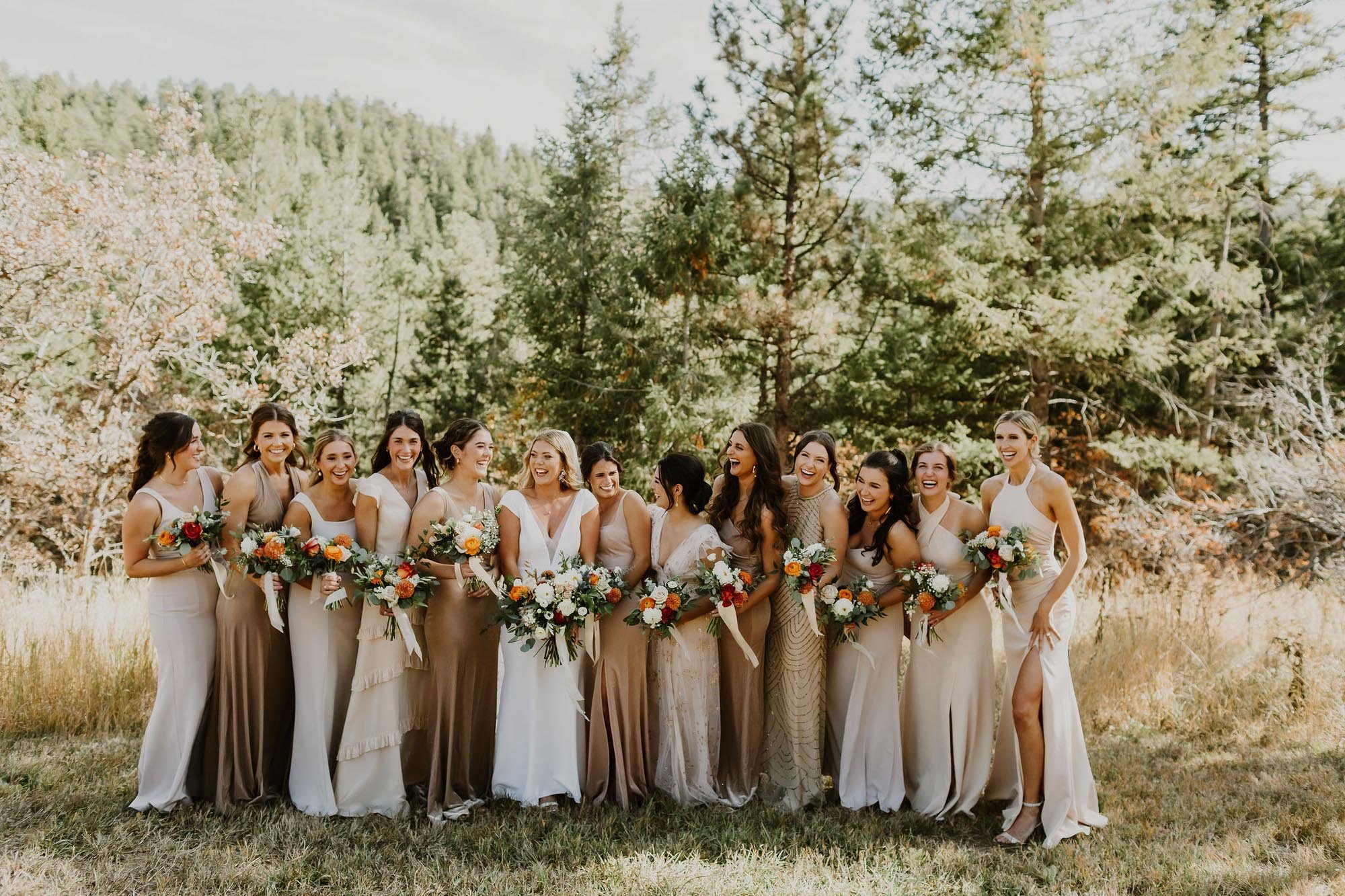  a wedding party all laughing and smiling for a bride’s big day. the bridesmaids are wearing various champagne colored dresses and have bouquets with orange, white, and rust colored flowers. they are standing on a mountain in colorado with trees in t