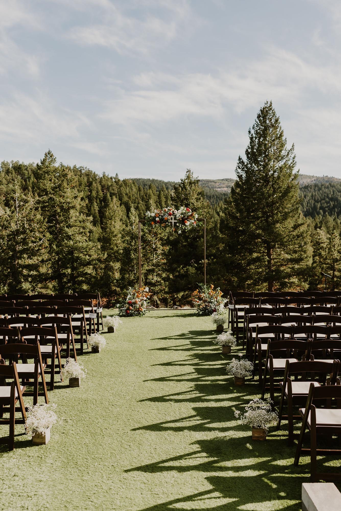  A lovely floral ceremony backdrop with florals in orange, rust and white sits perched on a hillside surrounded by evergreen trees. The aisles are lined with dark wooden folding chairs and floral arrangements made of babies breath. 