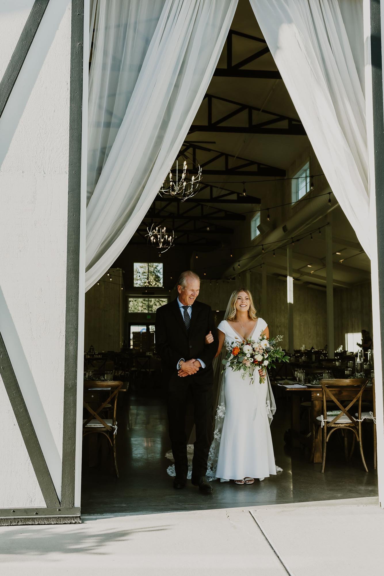  A bride smiling getting ready to walk down the aisle with her father. They are standing in the doorway of a lovely barn wedding venue and the bride is holding a colorful floral bouquet 