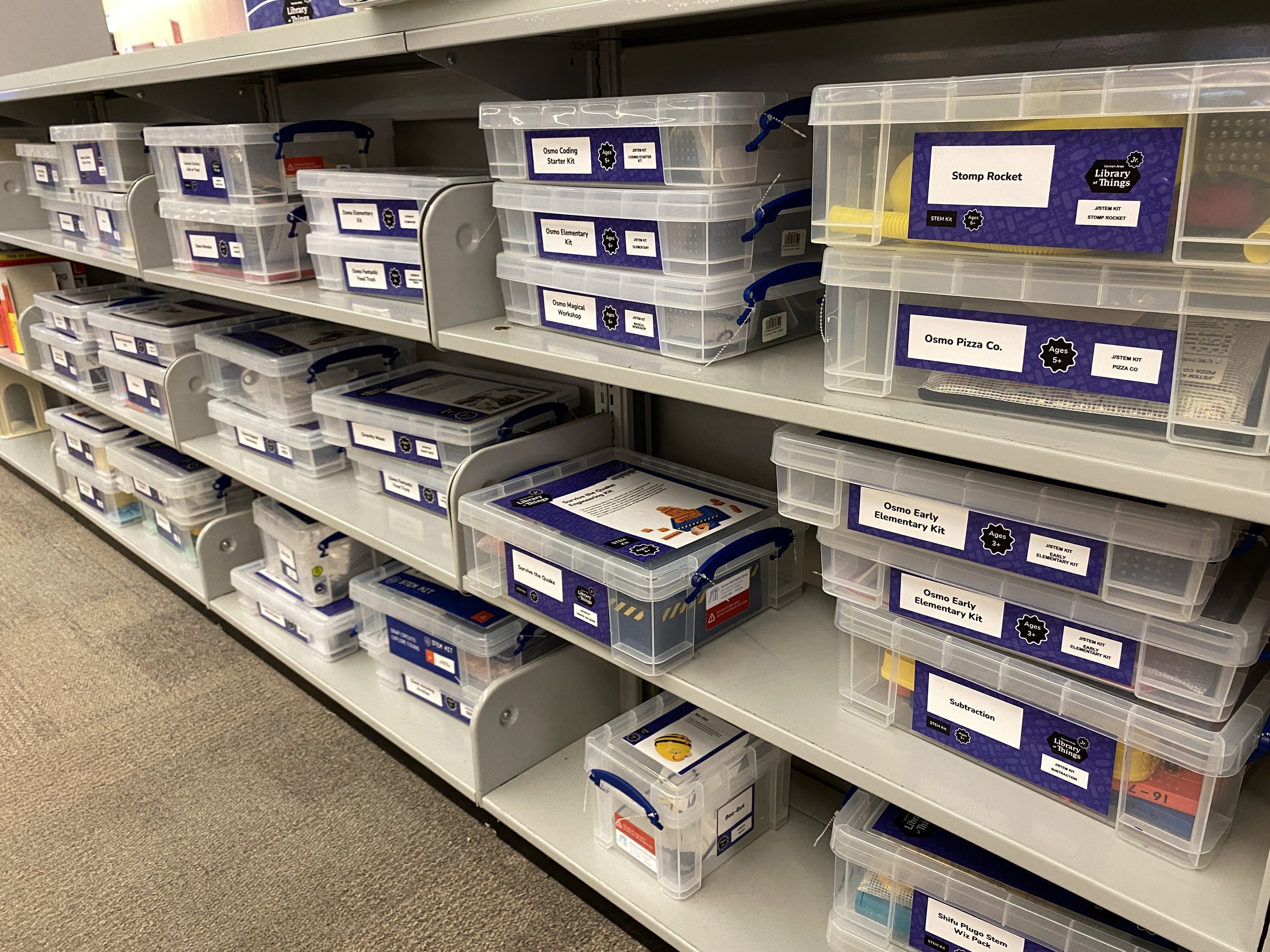 Library shelves holding rows of clear plastic bins with blue and white labels identifying youth STEM kit lending items such as Osmo kits, Stomp Rocket, and Subtraction games.