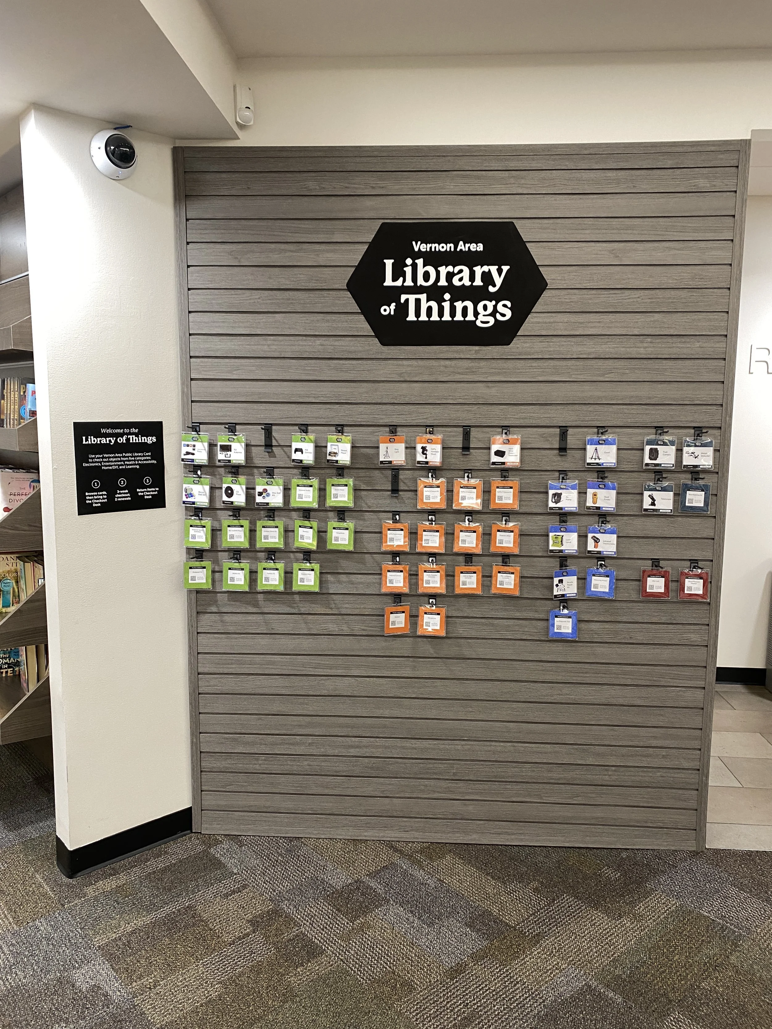 A slatwall display in the Vernon Area Library of Things, showing rows of color-coded hanging cards organized by category beneath a large black hexagonal logo.